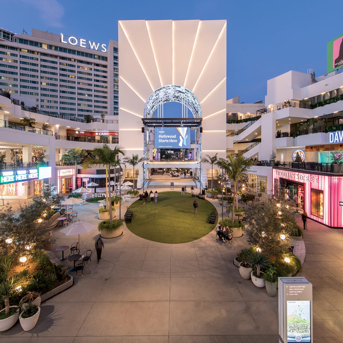 Aerial twilight view of Ovation Hollywood open-air shopping center courtyard with illuminated LED globe arch, "Hollywood Starts Here" digital screen, Loews Hotel, Victoria's Secret, and landscaped central lawn
