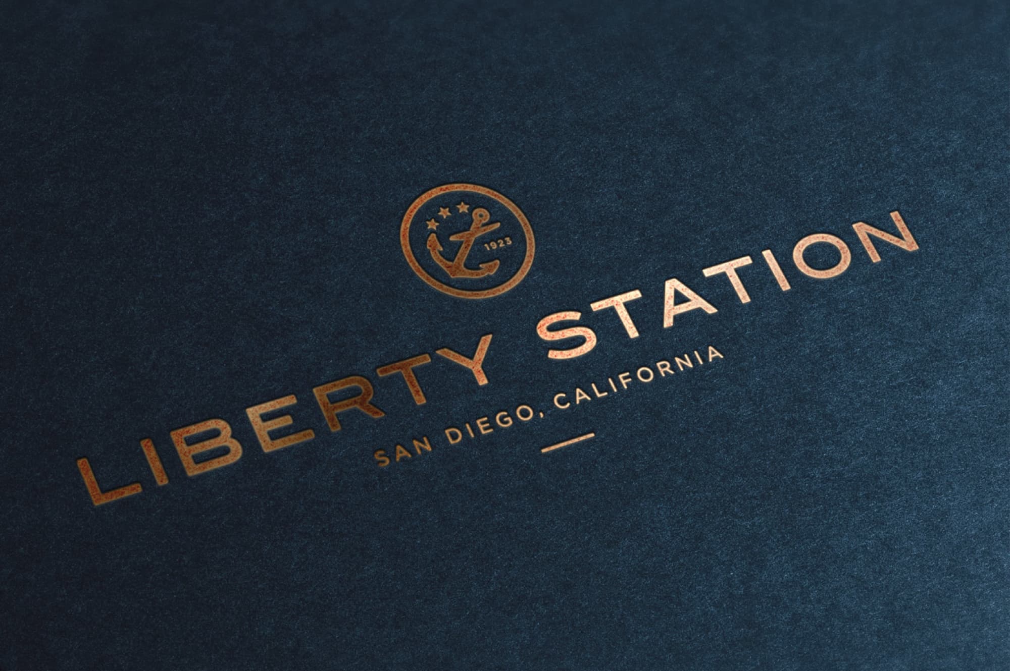 Copper foil-stamped 'Liberty Station, San Diego, California' logo with an anchor and three stars emblem dated 1923, on a textured navy blue surface.