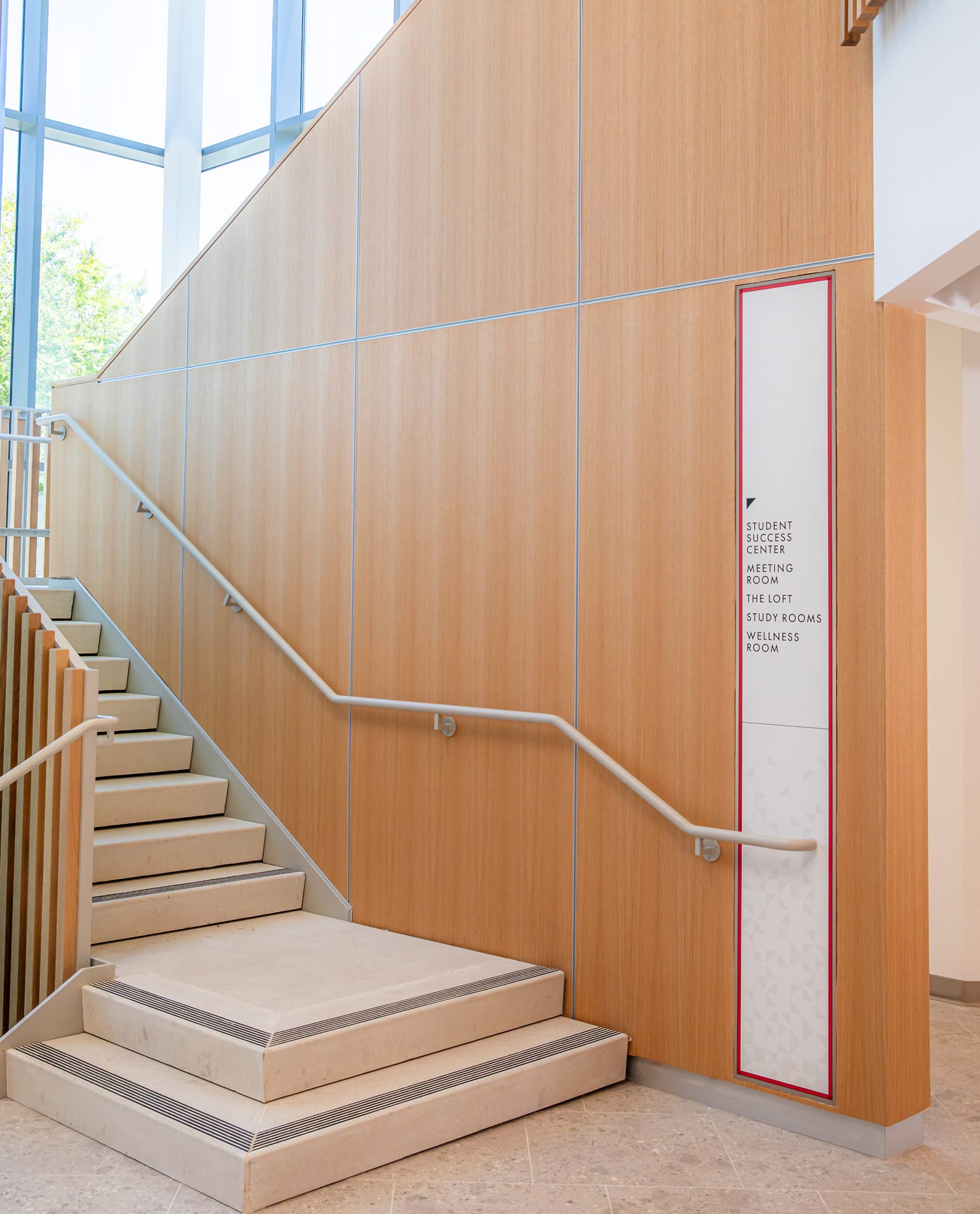 RSM Design Chapman University Rinker Campus Center Irvine — directional wayfinding signage panel with Chapman red border detail mounted on wood wall beside light-filled staircase, directing to Student Success Center, study rooms, and wellness room