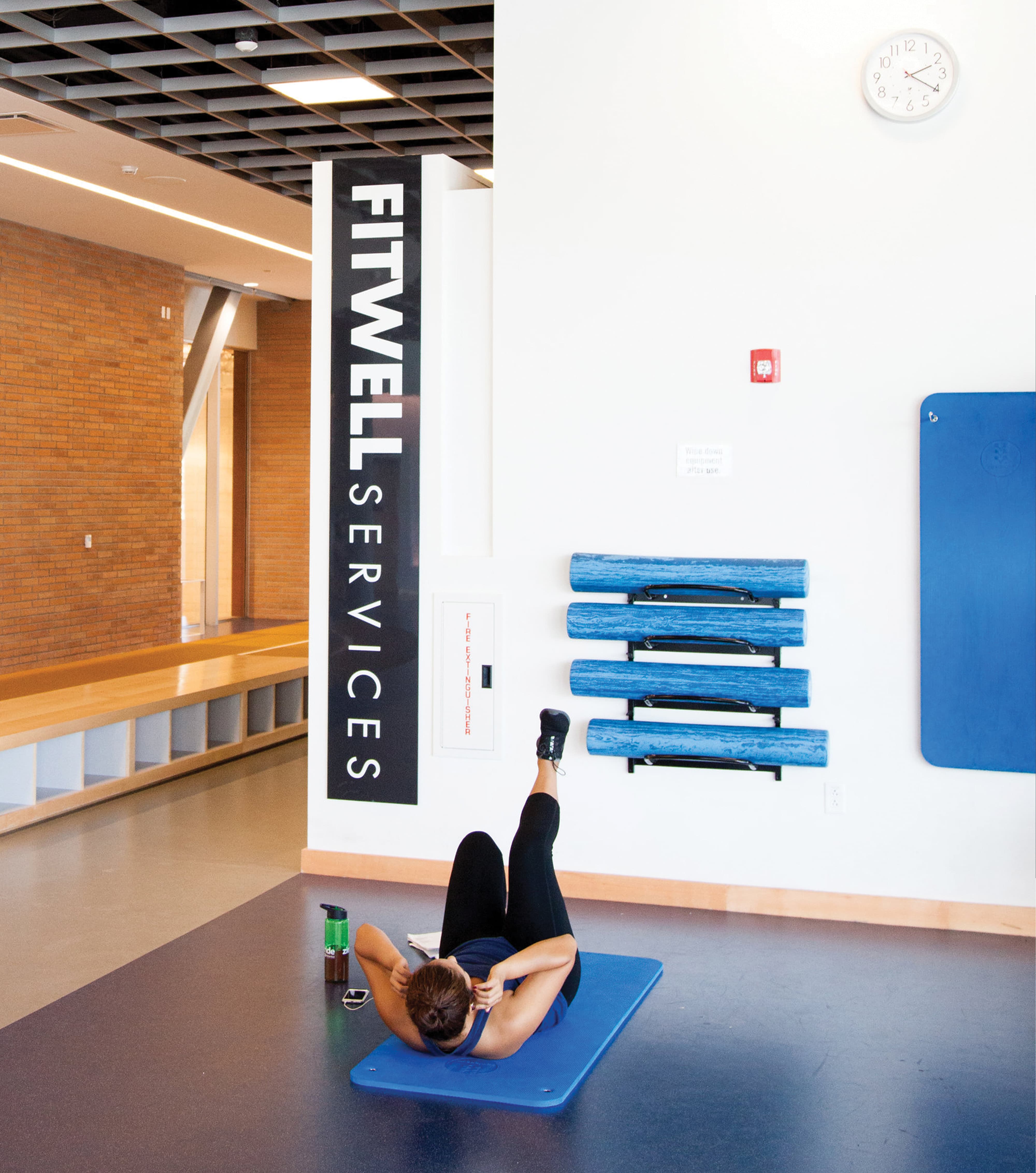 Woman doing an ab exercise on a blue mat in a gym area beneath a vertical 'Fitwell Services' sign, with foam rollers racked on the wall and a stretching mat nearby.