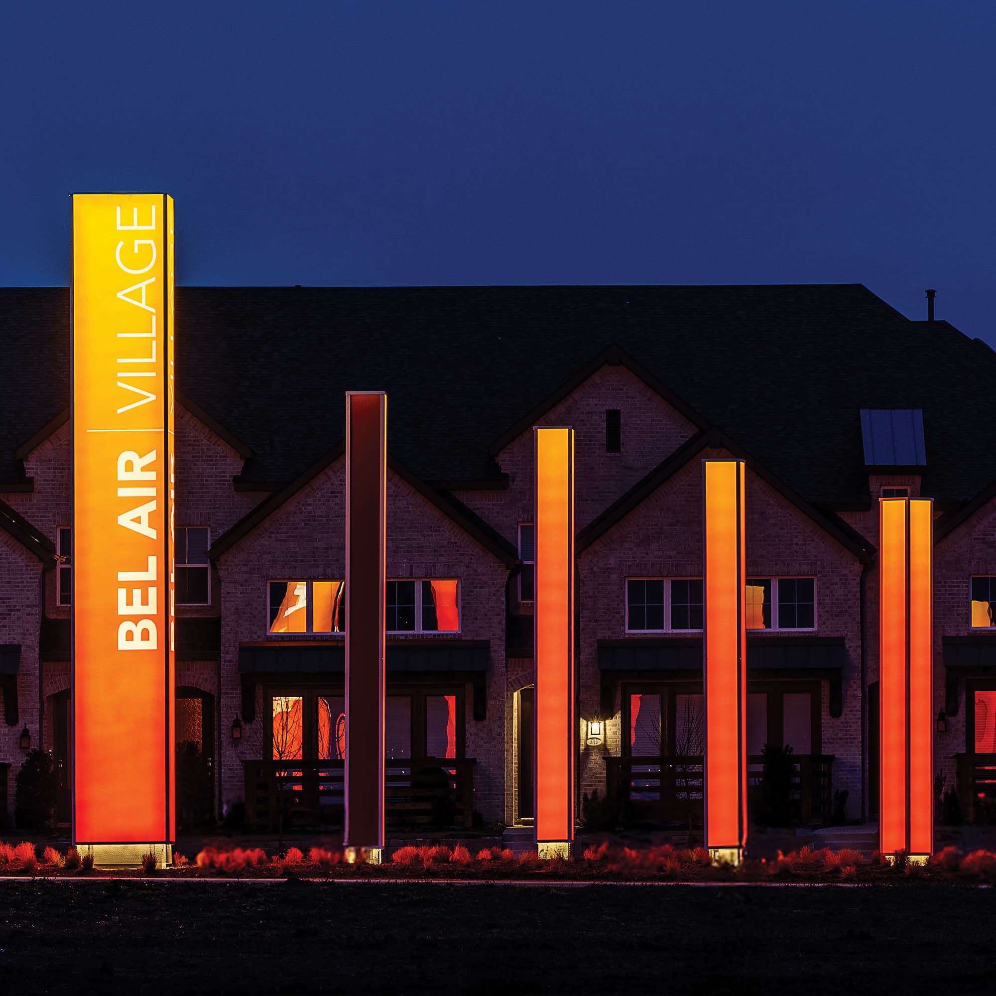 A row of five pylons stand in front of a large, brick, 2.5-story, multifamily home at nighttime. The leftmost pylon has text reading “Bel Air Village”, rotated 270-degrees and running vertically from bottom to top. All five pylons are illuminated with an yellow-to-orange gradient.