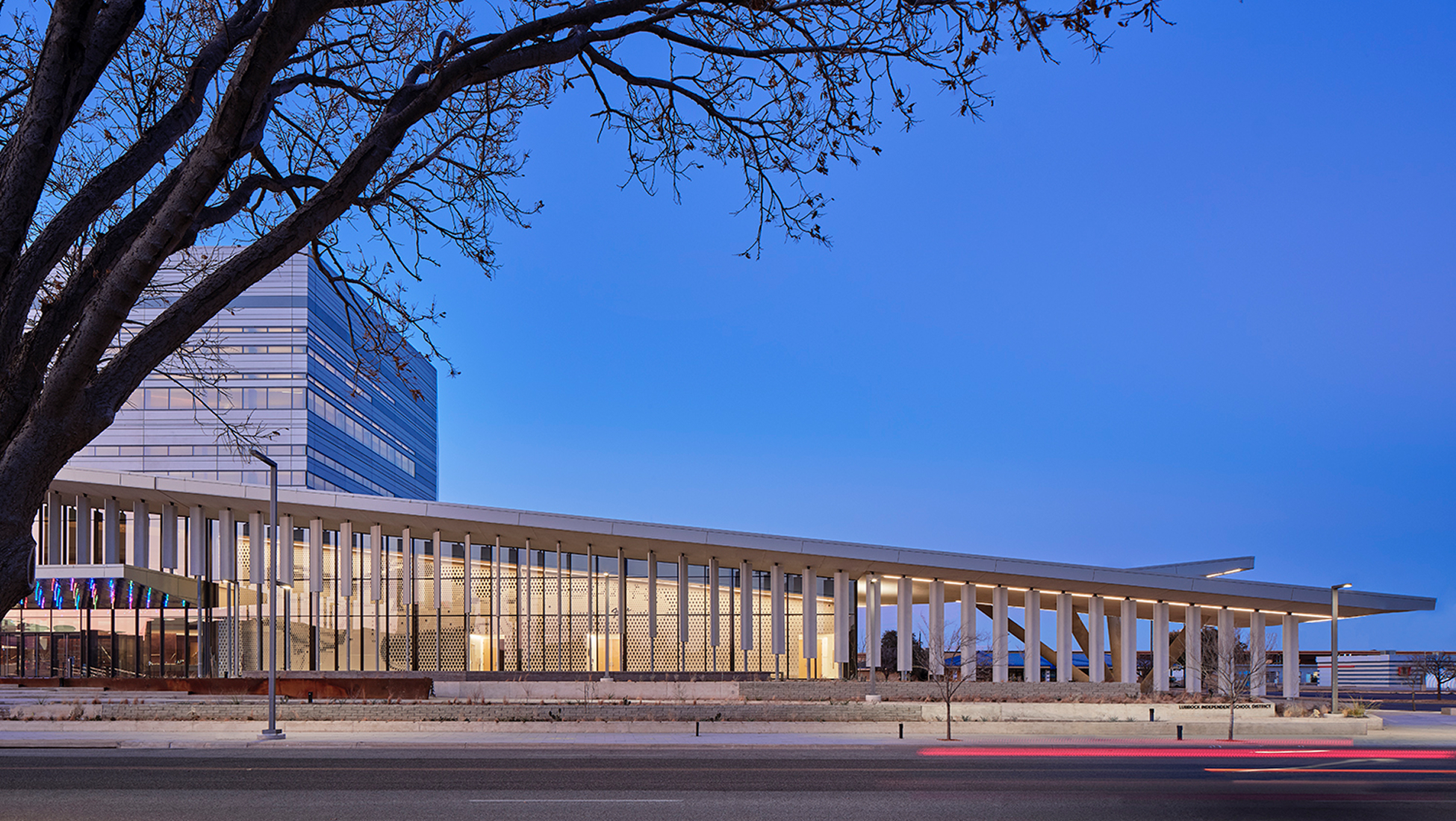 Modern Lubbock Independent School District administrative building at dusk, featuring a glass curtain wall facade with white columns, illuminated interior, and a angular multi-story tower, Lubbock Texas