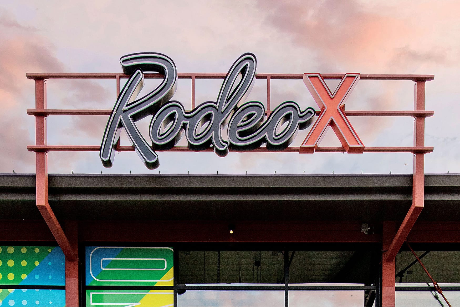 Rooftop sign reading 'Rodeo X' in script lettering mounted on a red steel frame above a building with colorful geometric window graphics, set against a pink and gray sunset sky.