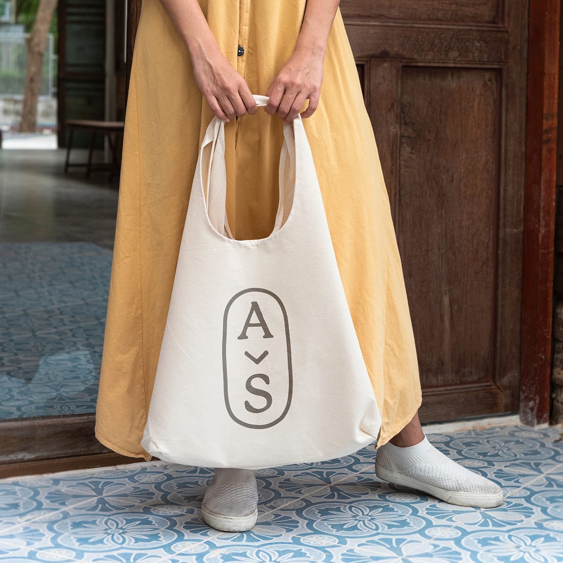 Woman in yellow dress holding canvas tote bag printed with Avenue South monogram over patterned tile floor, brand identity by RSM Design.