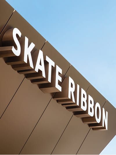 Skate Ribbon facility sign at Gene Leahy Mall in downtown Omaha, Nebraska, featuring bold white dimensional lettering on a modern dark metal canopy facade against a clear blue sky