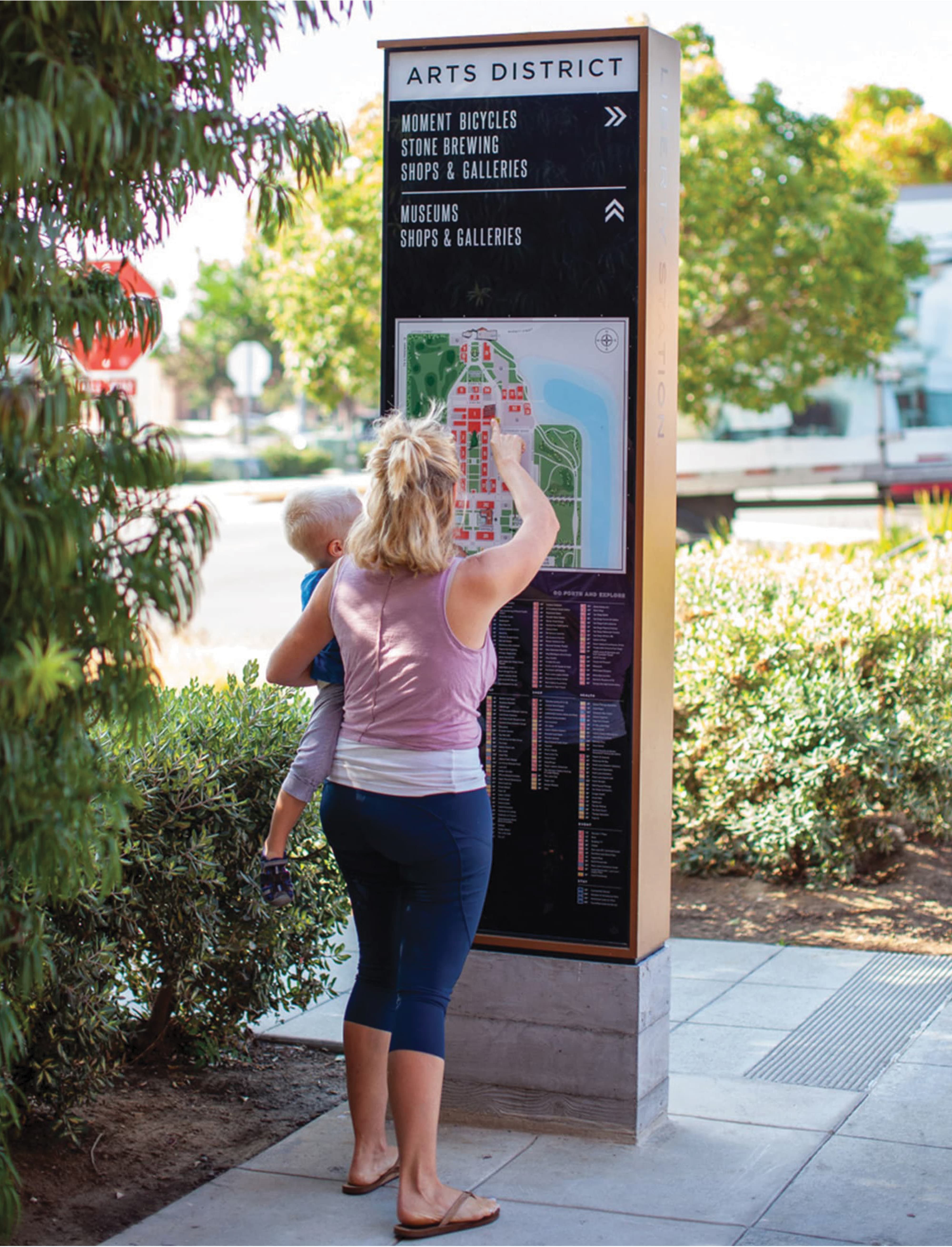 Woman holding a toddler pointing at a color-coded site map on a Liberty Station wayfinding pylon reading 'Arts District, Moment Bicycles, Stone Brewing, Shops & Galleries, Museums,' set on a sidewalk lined with trees and shrubs.