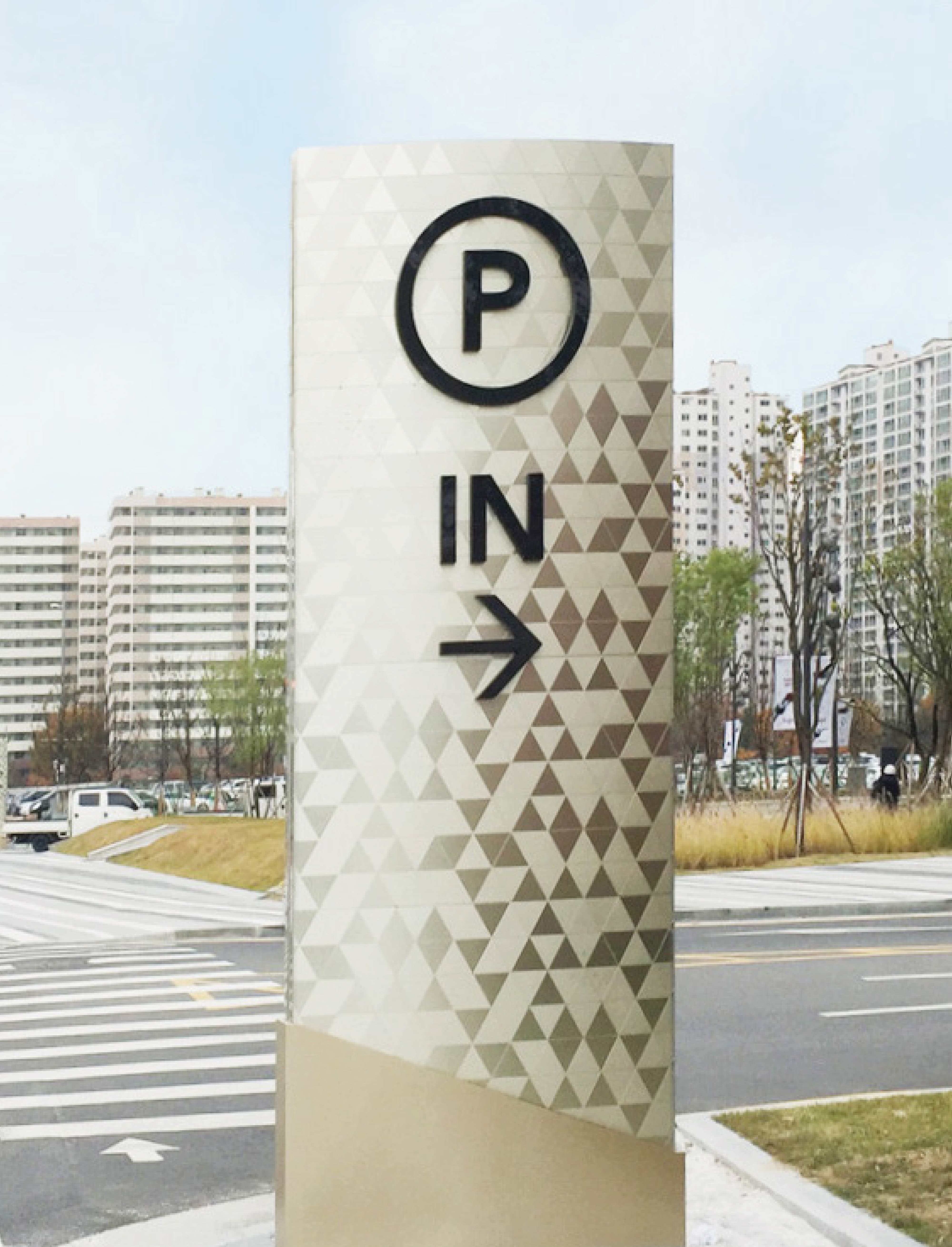Tall triangular-patterned parking wayfinding pylon with a 'P' symbol, 'IN' text, and right-pointing arrow, standing at a street intersection with high-rise apartment buildings in the background.