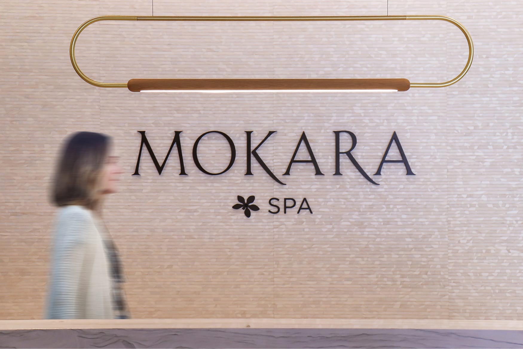 Mokara Spa interior lobby signage at Omni PGA Frisco featuring dimensional letters on textured stone wall, brass oval pendant light, and blurred guest walking in foreground