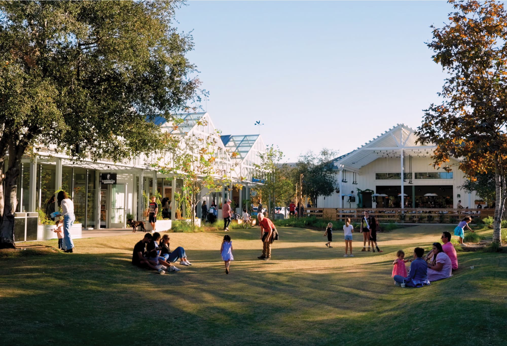Families and visitors gathering on the central lawn at River Street Marketplace in San Juan Capistrano at golden hour, surrounded by white barn-style retail and greenhouse structures amid mature trees, showcasing placemaking and experiential design for a mixed-use waterfront retail environment by RSM Design.