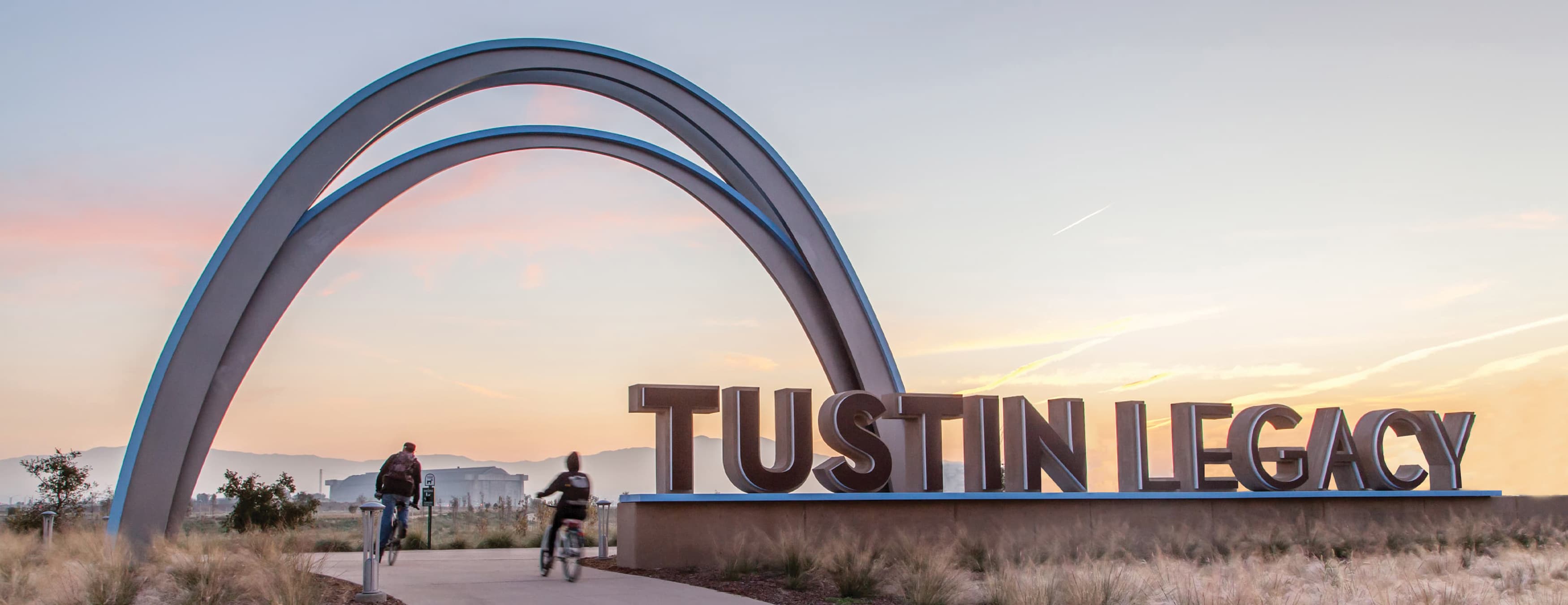 Large Tustin Legacy logo and 30 foot arch logo mark as gateway entry over pedestrian trail with two people on a bike ride