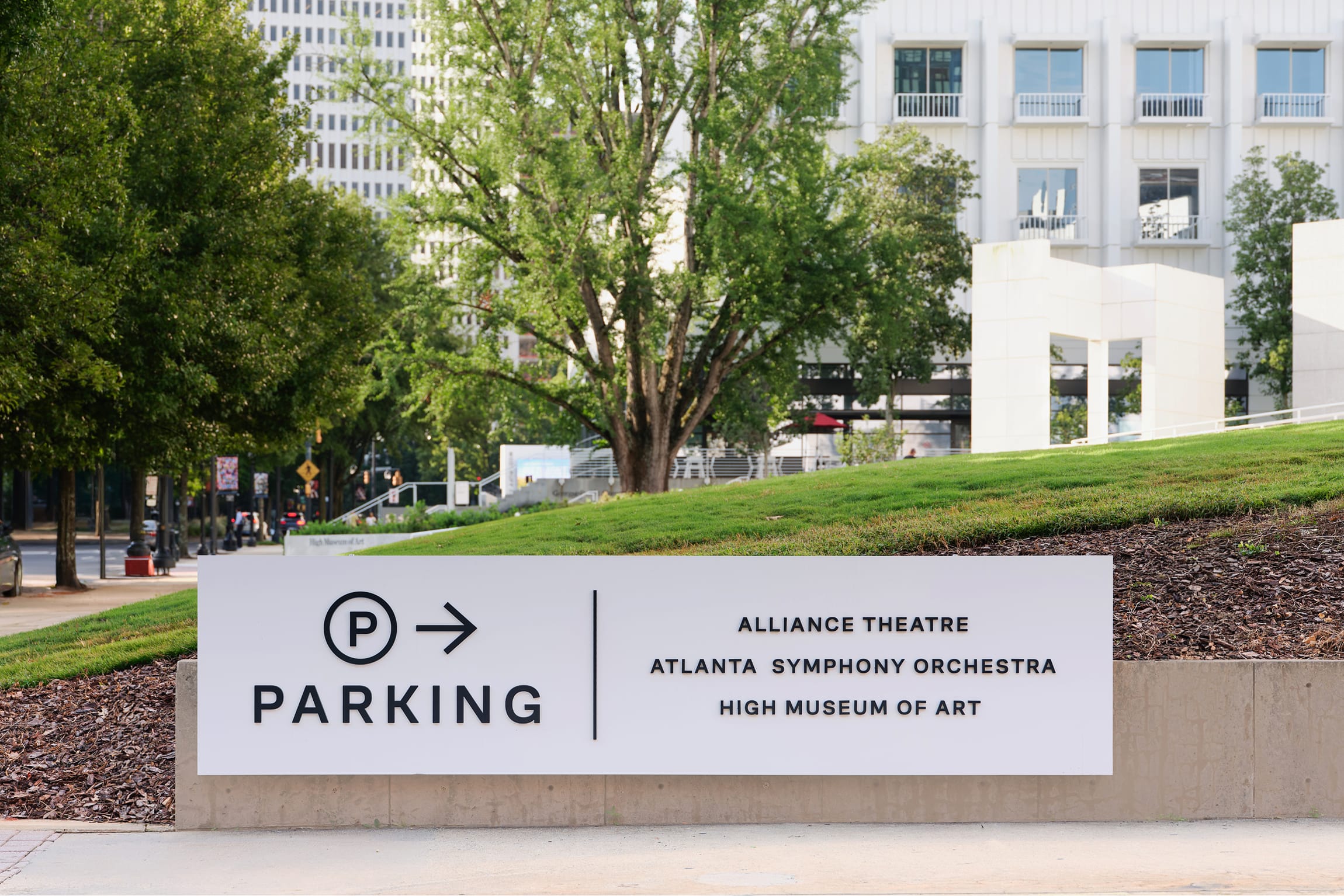 Ground-level directional sign with a "P" parking icon and arrow, listing the Alliance Theatre, Atlanta Symphony Orchestra, and High Museum of Art, set on a landscaped berm with the campus visible behind – campus wayfinding and environmental graphic design by RSM Design.