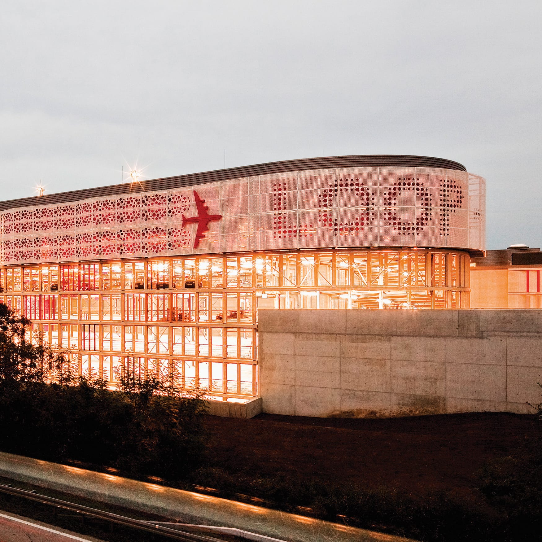 Modern architectural building exterior at sunset featuring a curved perforated facade with graphic patterns, illuminated glass curtain walls, structural steel detailing, and warm glowing interior light.