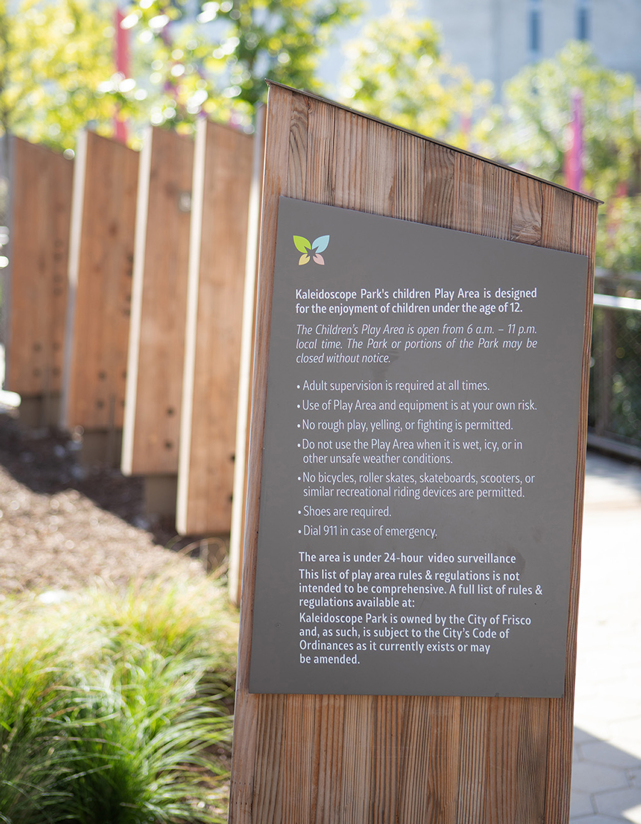 Kaleidoscope Park children's play area rules and regulations sign on cedar wood post kiosk with colorful leaf logo, City of Frisco public park signage, Frisco, Texas