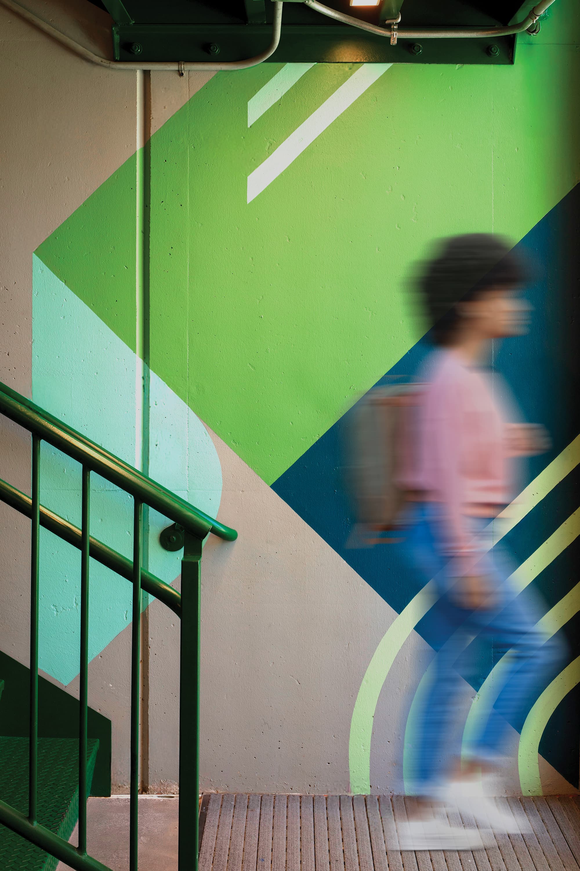 Motion-blurred person walking through a stairwell painted with a bold green, teal, and navy geometric mural, beside a dark green metal handrail.