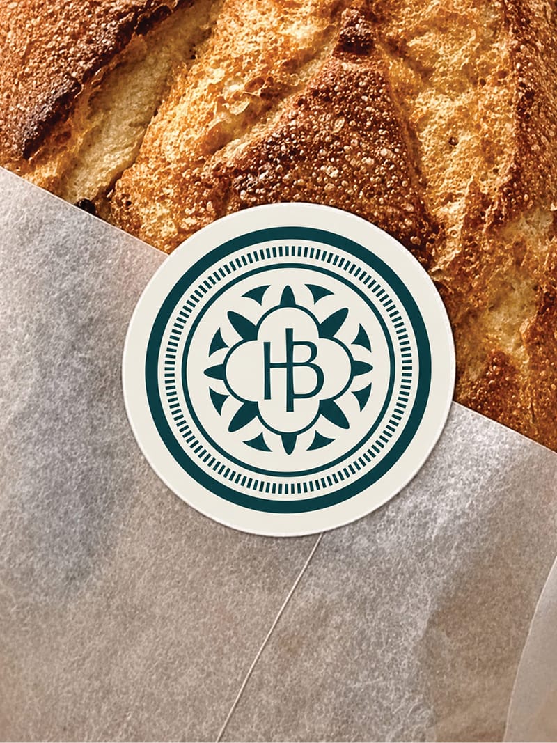 Round "HB" sticker with ornate mandala-style border on wax paper over crusty sourdough bread — brand identity design by RSM Design