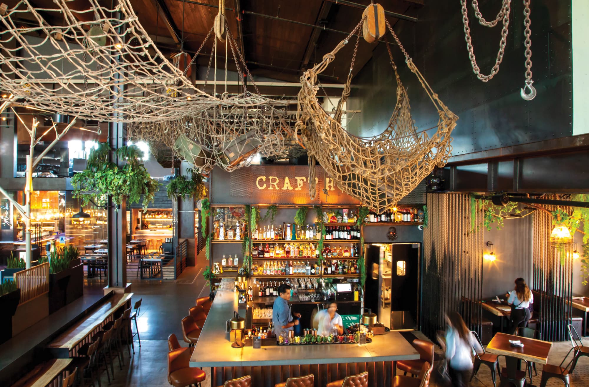 Interior of Crafthouse restaurant and bar featuring a dramatic industrial-nautical themed space with suspended rope netting, heavy chain rigging, lush hanging greenery, and a backlit full-service bar beneath a dimensional metal letter logo — illustrating RSM Design's experiential graphics and branded environments, placemaking, and specialty graphics services within the food and beverage and mixed-use markets