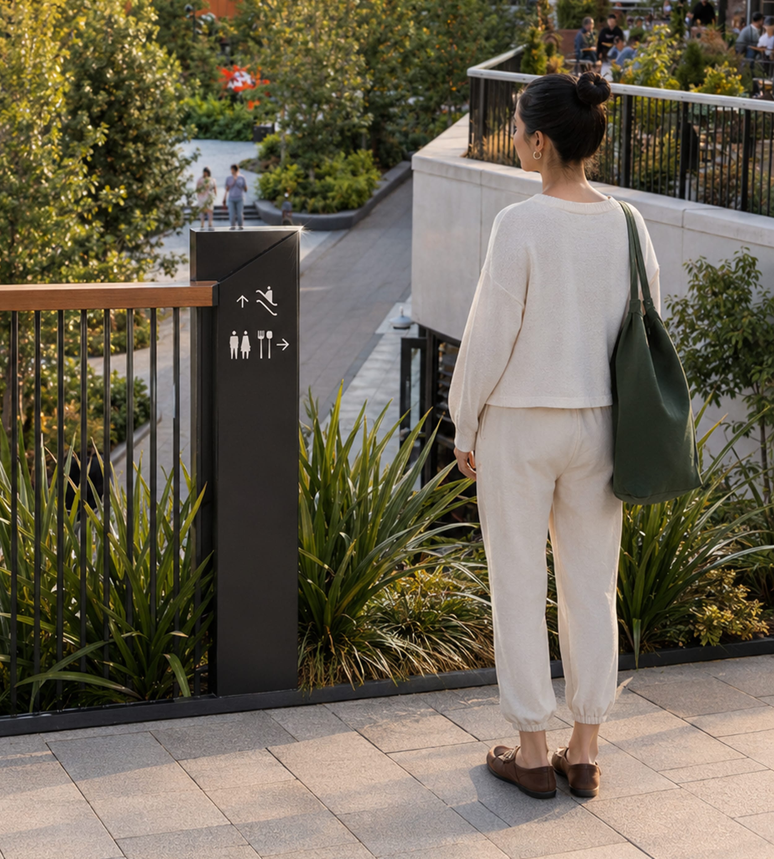 Woman standing on an outdoor terrace beside a black pylon sign with pictograms for escalator, restrooms, and dining, landscaped plaza visible below — wayfinding design by RSM Design