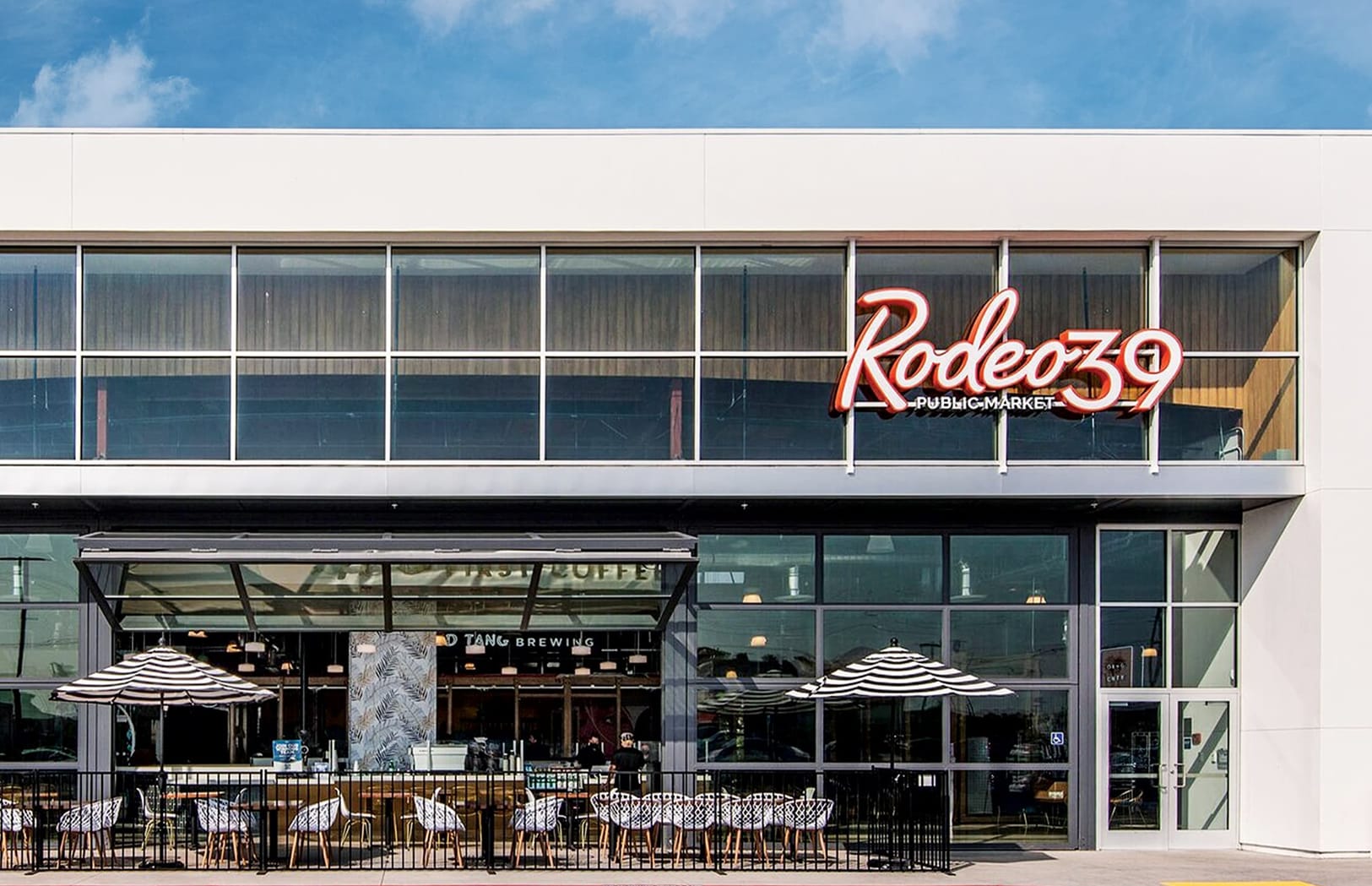 Daytime exterior facade of Rodeo 39 Public Market in Stanton, CA, featuring a bold red neon sign above floor-to-ceiling storefront glazing, with an outdoor patio below shaded by black-and-white striped umbrellas and seating for Old Tang Brewing.
