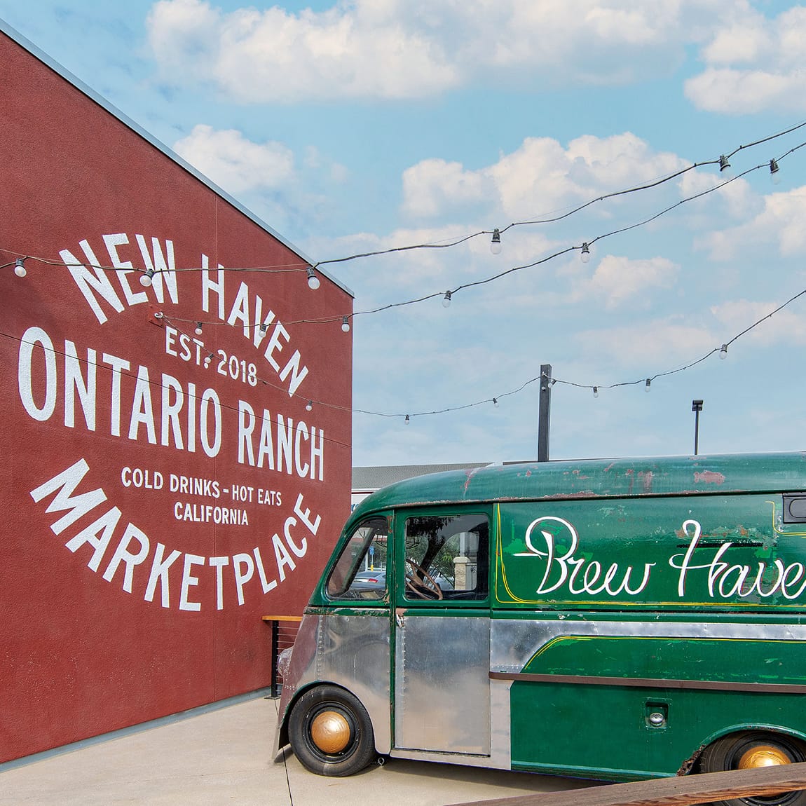 Vintage green 'Brew Haven' delivery van parked beside a red building painted with a circular 'New Haven Ontario Ranch Marketplace, Est. 2018, Cold Drinks - Hot Eats, California' mural, strung with string lights against a blue sky.