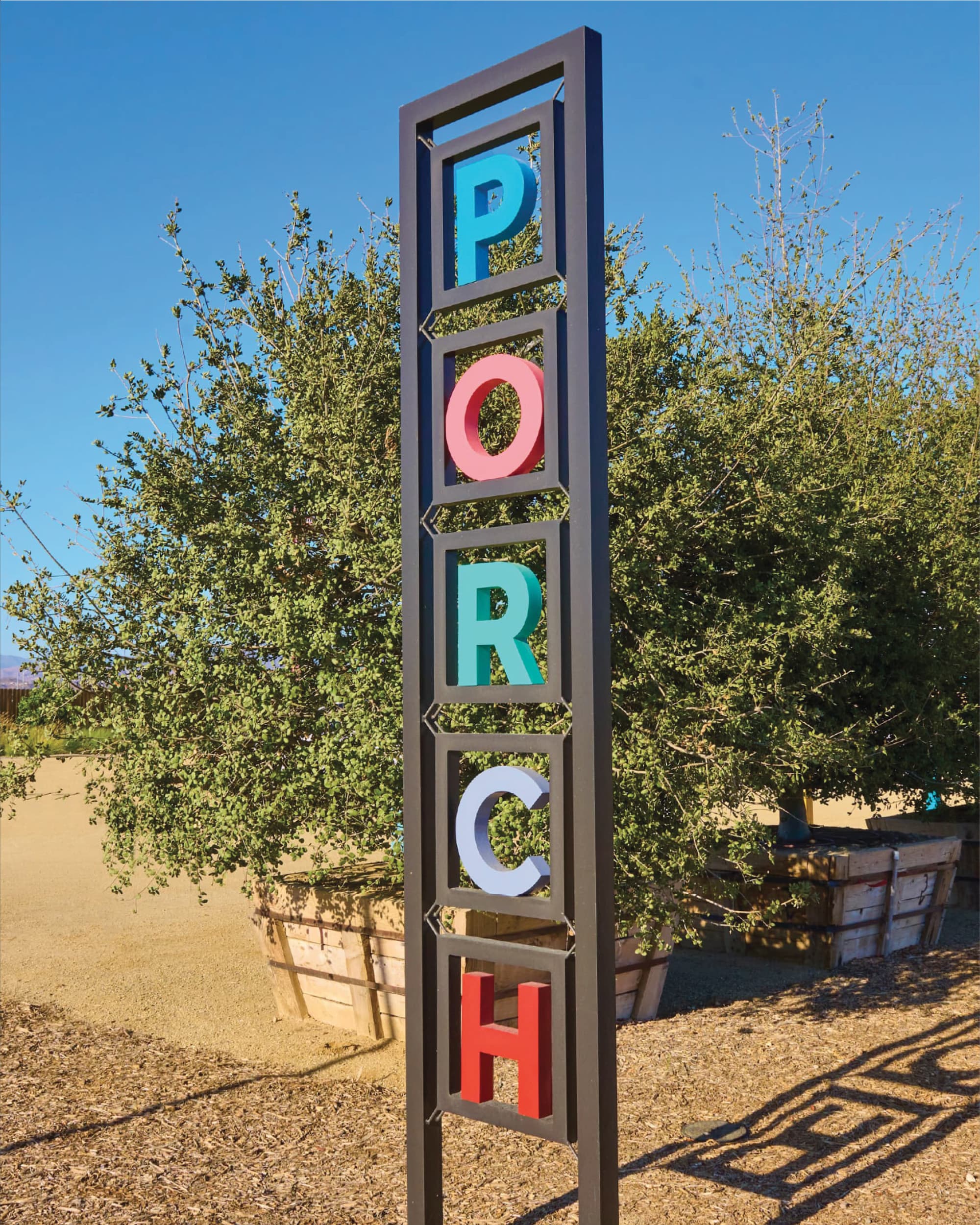 Tall vertical sign spelling 'PORCH' in multicolored 3D letters set within a dark metal frame, standing in front of a large shrub and raised wooden garden beds under a clear blue sky.