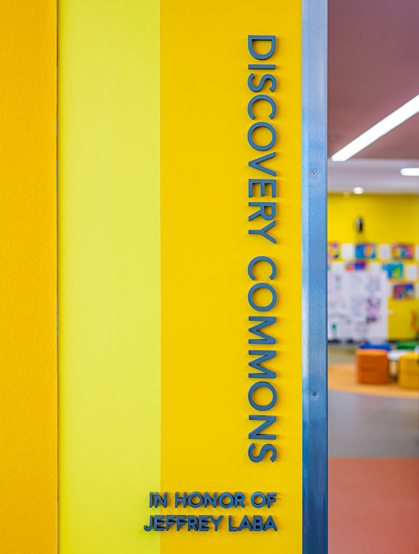 Bright yellow entrance wall with vertical blue dimensional lettering reading 'Discovery Commons, in honor of Jeffrey Laba,' beside a doorway into a colorful classroom with student artwork and orange and yellow seating cubes.