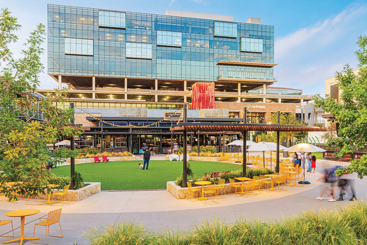 People gathering at an activated public lawn and outdoor plaza at Toyota Music Factory mixed-use entertainment district in Irving, Texas, featuring tenant identification signage, branded environmental graphics, and specialty art elements — RSM Design experiential placemaking and environmental graphic design for mixed-use, food and beverage, workplace, and streetscape destinations.
