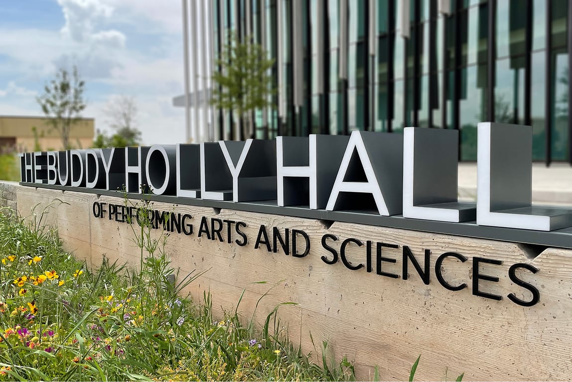 Exterior monument sign for The Buddy Holly Hall of Performing Arts and Sciences featuring large dimensional letters on a concrete base surrounded by native wildflowers, with the glass and steel building facade visible in the background, Lubbock Texas