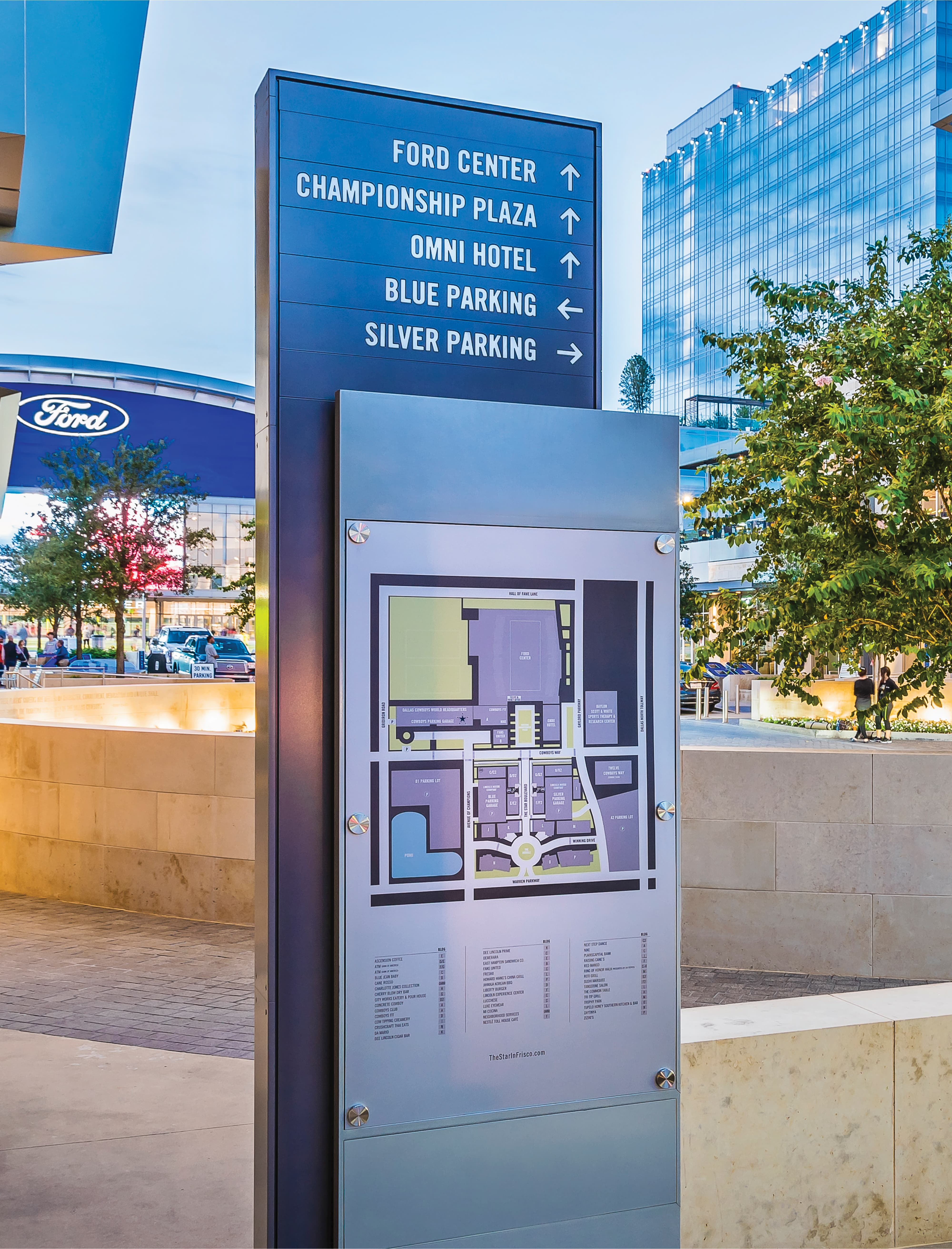 A two-part exterior wayfinding and directory sign post at The Star in Frisco featuring a navy blue directional blade with white arrow-led destinations to Ford Center, Championship Plaza, Omni Hotel, Blue Parking, and Silver Parking, above a site map panel with color-coded district plan and tenant directory listing, set against the Ford Center arena and a glass tower at dusk — showcasing RSM Design's wayfinding and signage design, master sign programs, and environmental graphic design services applied within the sports and entertainment and mixed-use markets