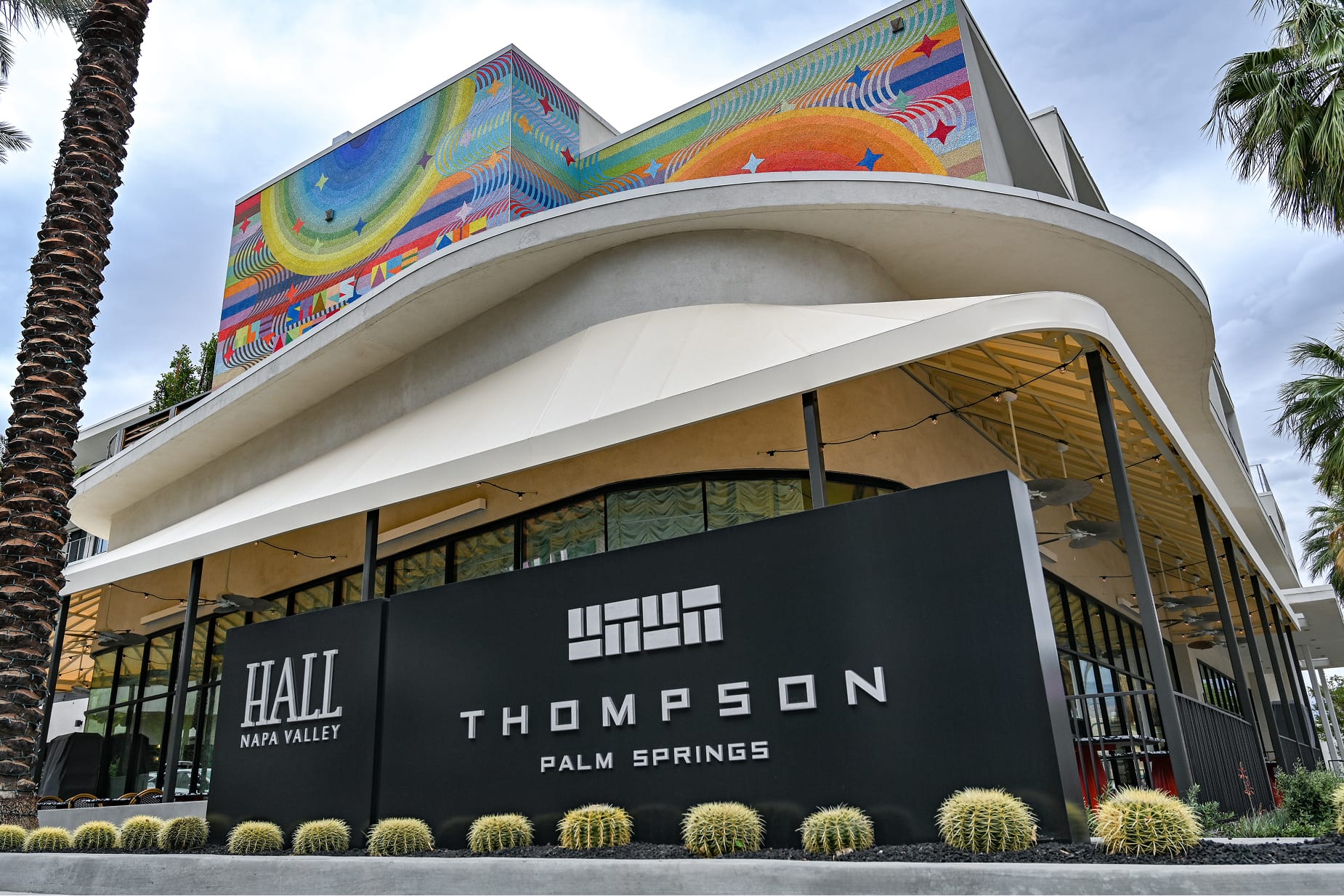 Exterior facade of Thompson Palm Springs hotel featuring a sweeping curved roofline, a large-scale colorful mural wrapping the upper story, and a black monument sign for Hall Napa Valley and Thompson Palm Springs, flanked by golden barrel cacti and palm trees.