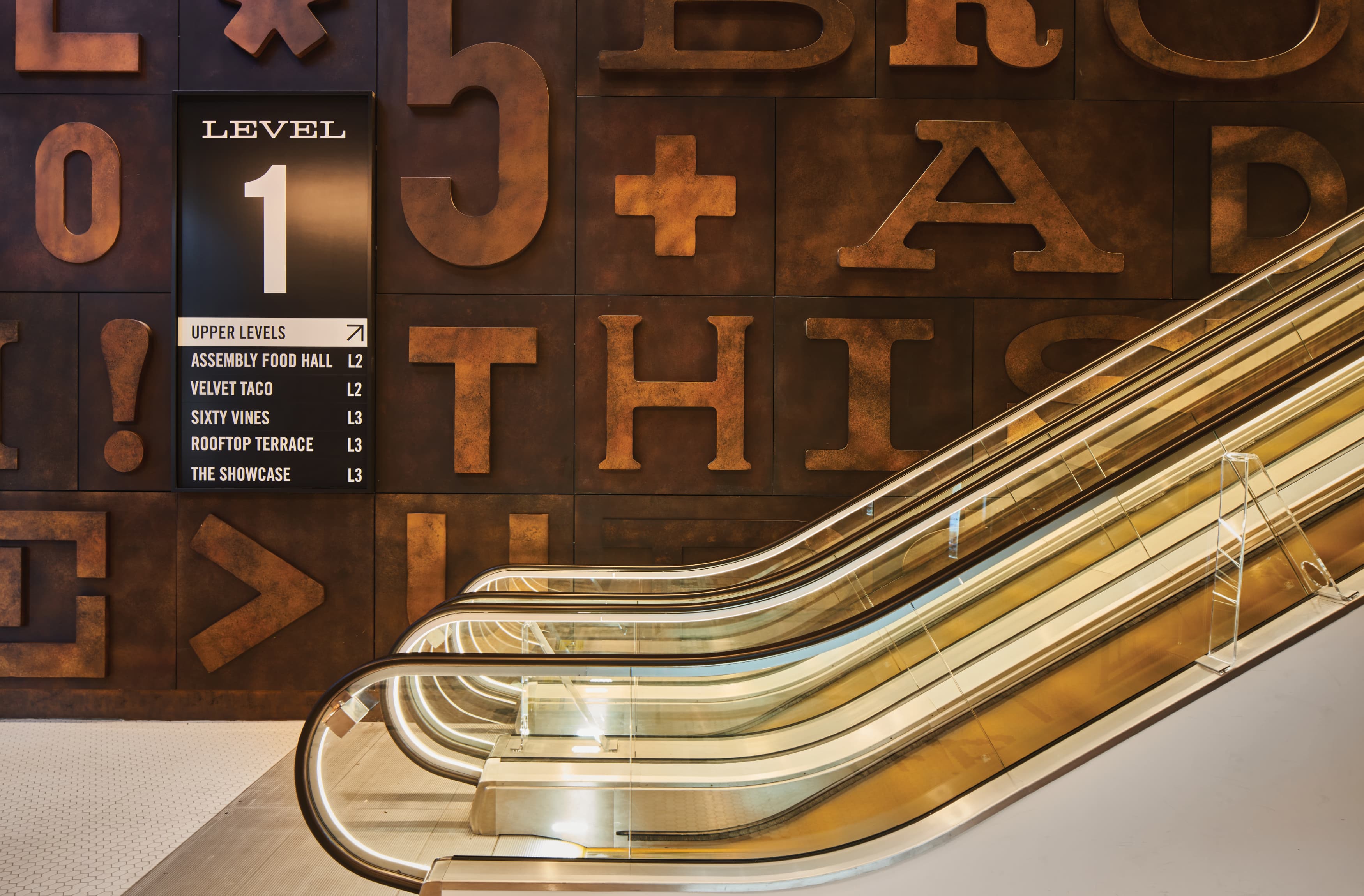 Image of the brown typographic wall with bold lettering differing in fonts and style in front of the escalators at Fifth + Broadway.