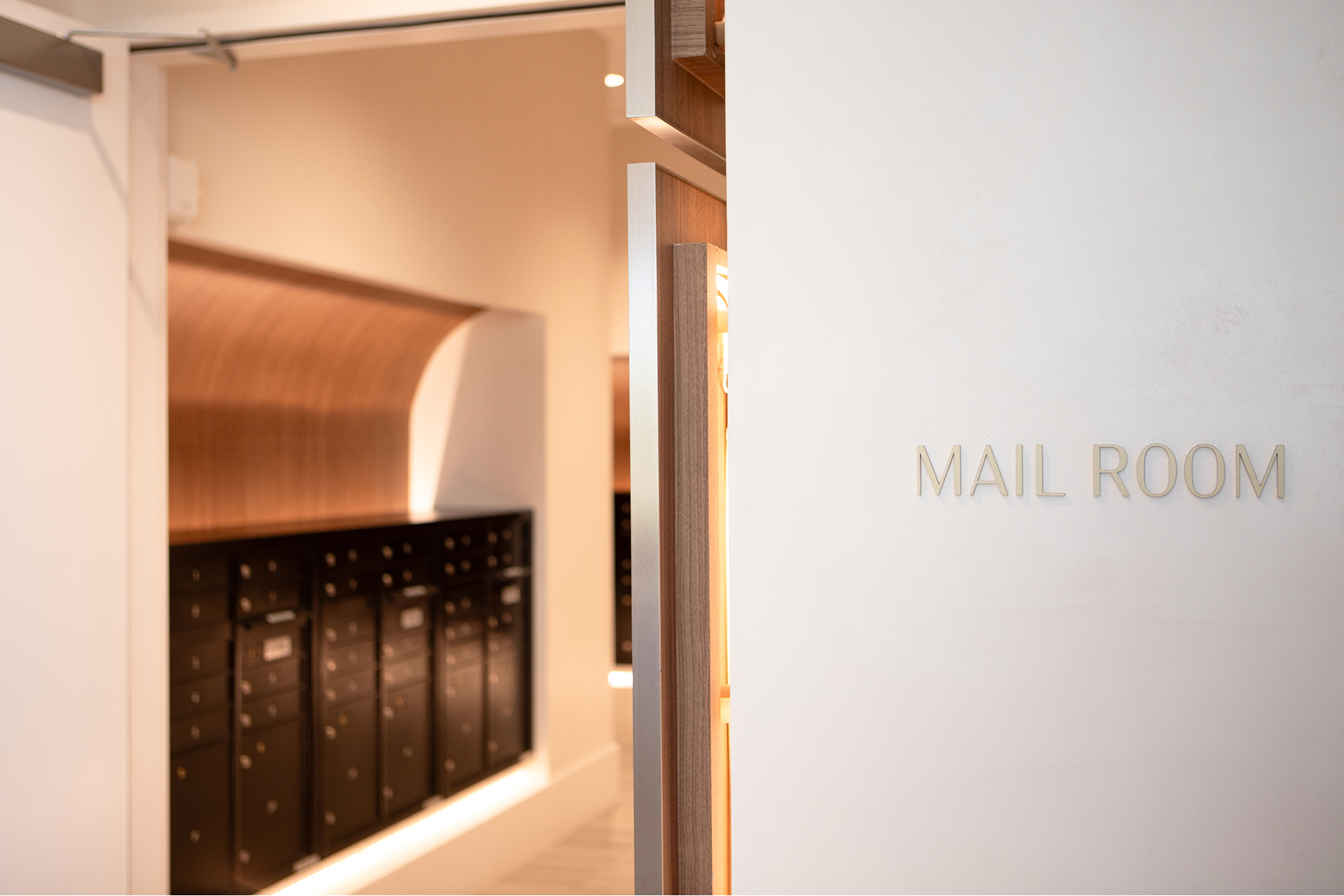 The Monarch at Hall Park interior wayfinding signage — dimensional brushed gold "Mail Room" room identification sign on white wall with warm wood and arched mailbox alcove interior, environmental graphic design by RSM Design