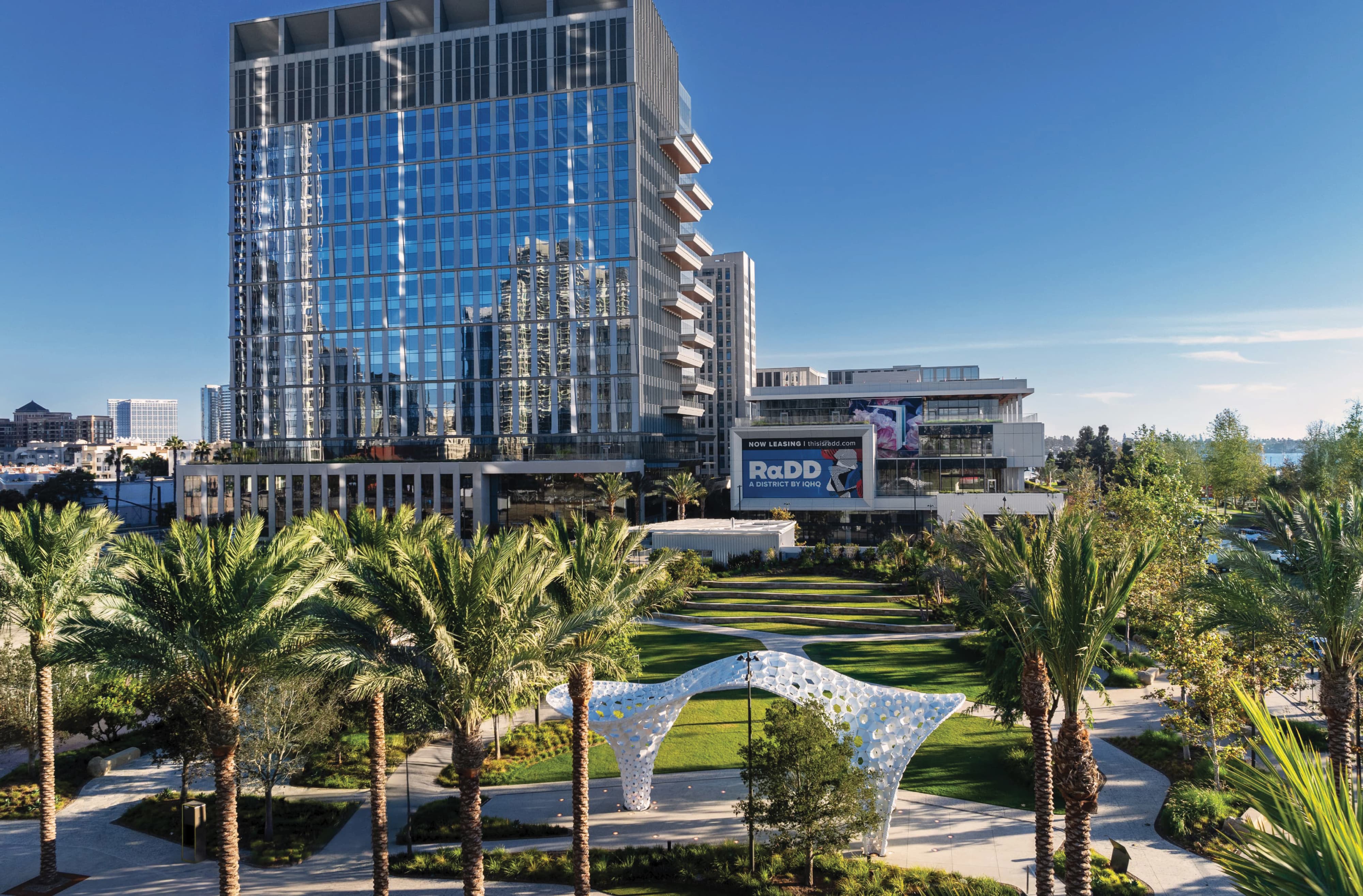 Elevated view of RαDD's waterfront district, showing a tall glass high-rise with a 'RαDD, A District by IQHQ, Now Leasing' banner, a terraced park with palm trees and a white perforated archway sculpture, and the San Diego bay visible in the distance.