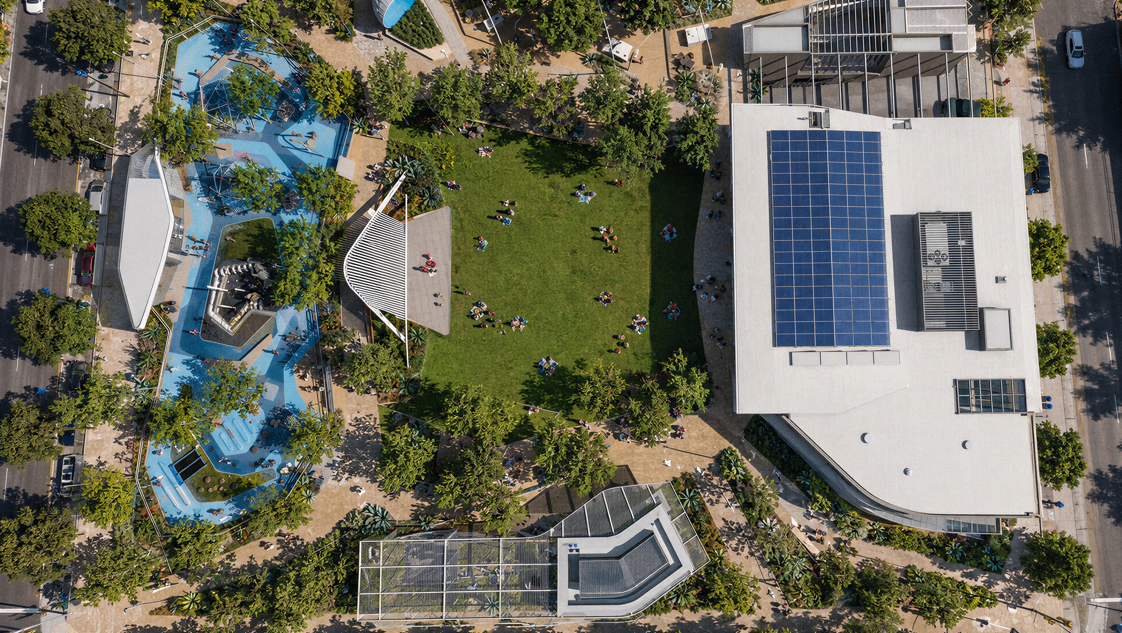 Aerial view of East Village Green park in San Diego, showing a central lawn with people relaxing, a blue splash pad and playground, a solar-paneled recreation center roof, a fenced sport court, and tree-lined pathways surrounded by city streets.