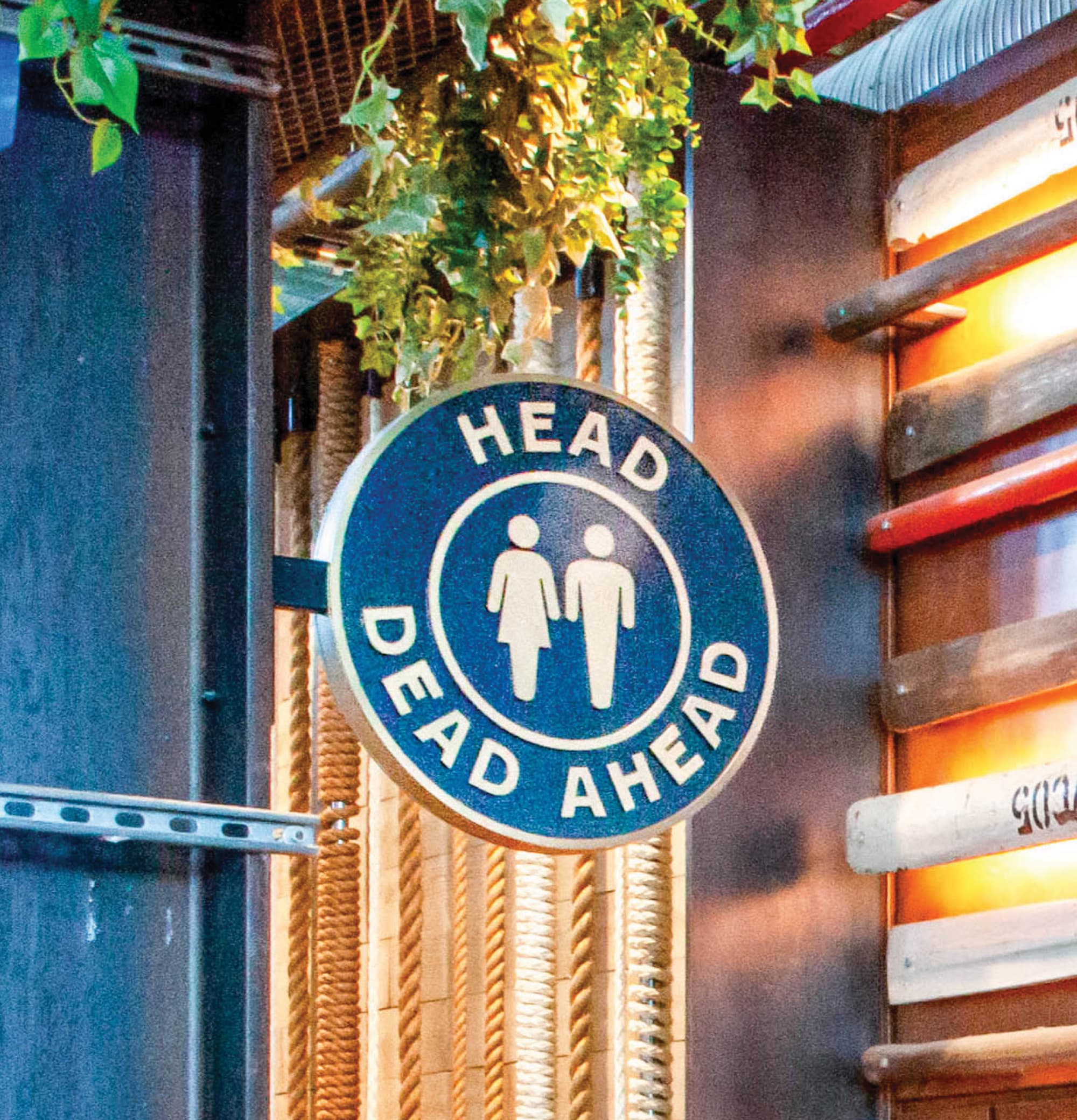 A circular navy blue projecting restroom sign reading "Head Dead Ahead" with universal gender pictograms, mounted among nautical rope and trailing ivy in an industrial-themed interior — showcasing RSM Design's playful and themed approach to wayfinding and signage design and specialty graphics within the food and beverage and mixed-use markets