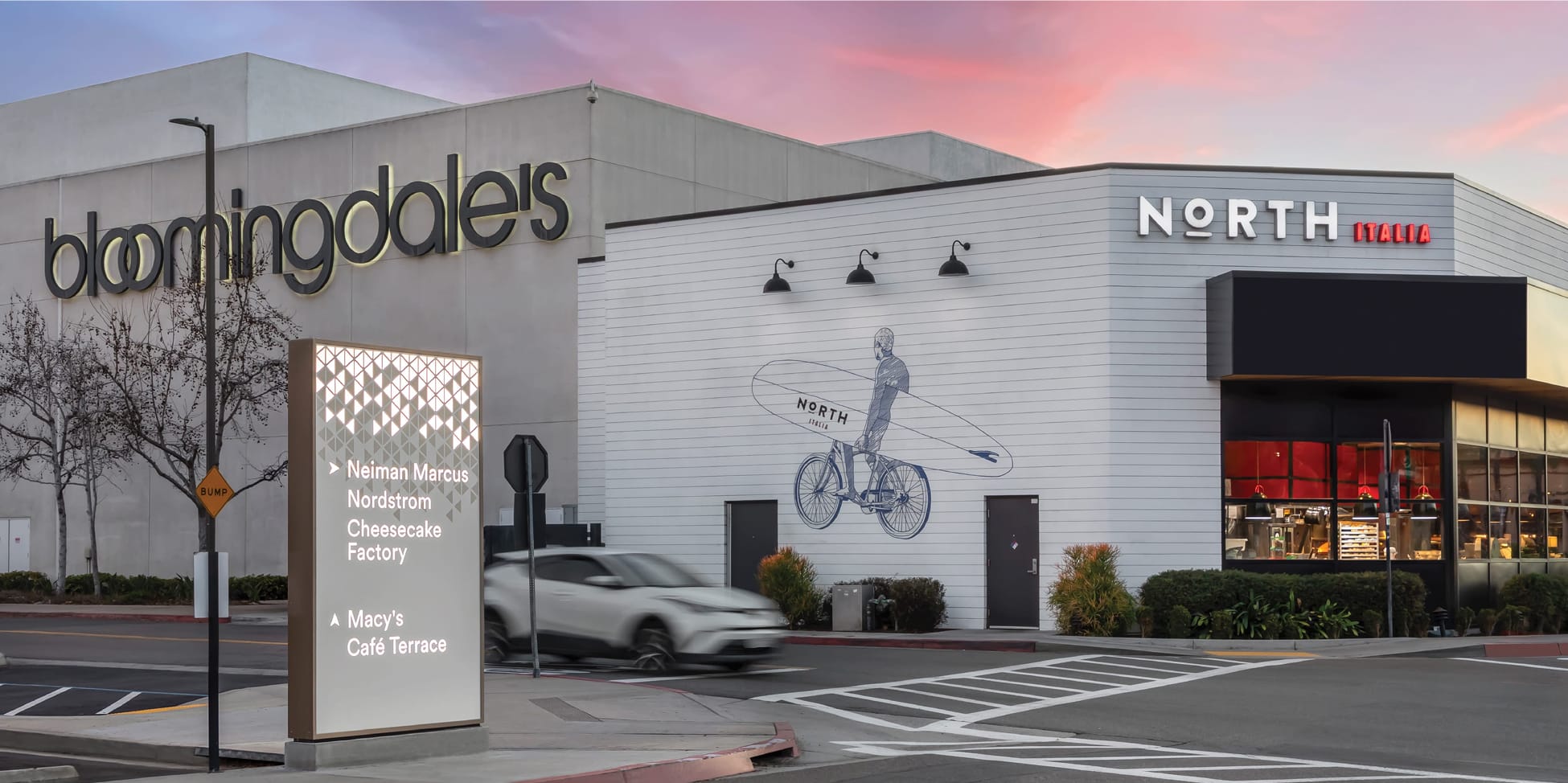 Exterior view of Fashion Valley Mall at sunset showing the Bloomingdale's anchor store, North Italia restaurant with a surfer mural on its facade, and an illuminated wayfinding pylon directing drivers to Neiman Marcus, Nordstrom, Cheesecake Factory, Macy's, and Café Terrace.