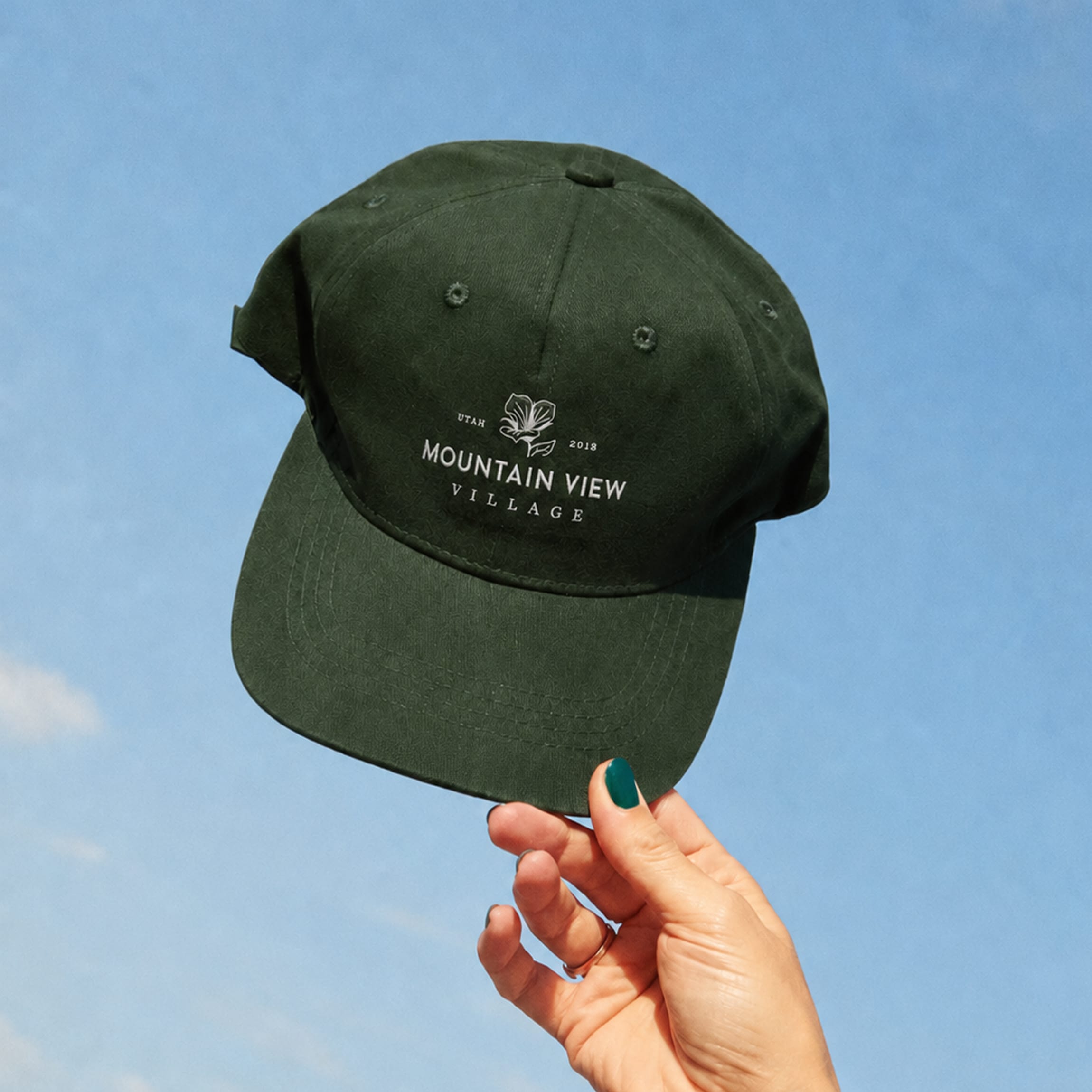 Hand holding a dark green baseball cap embroidered with the sego lily emblem and Mountain View Village wordmark against a blue sky — branding and environmental graphic design by RSM Design.