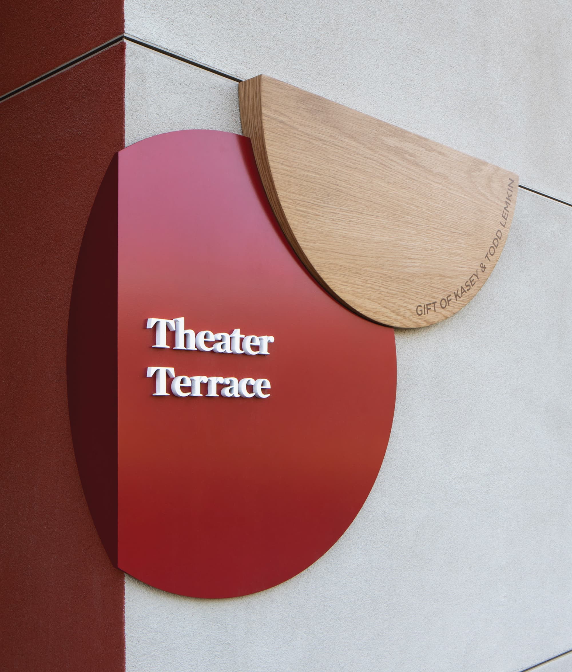 A sculptural donor recognition and space identification sign for "Theater Terrace" composed of a deep red oval panel with dimensional white letterforms overlapping a natural oak wood half-circle engraved with "Gift of Kasey & Todd Lemkin," mounted at the corner junction of a red and concrete exterior wall — showcasing RSM Design's donor recognition and specialty graphics services that artfully merge wayfinding, material craft, and philanthropic recognition within the education market