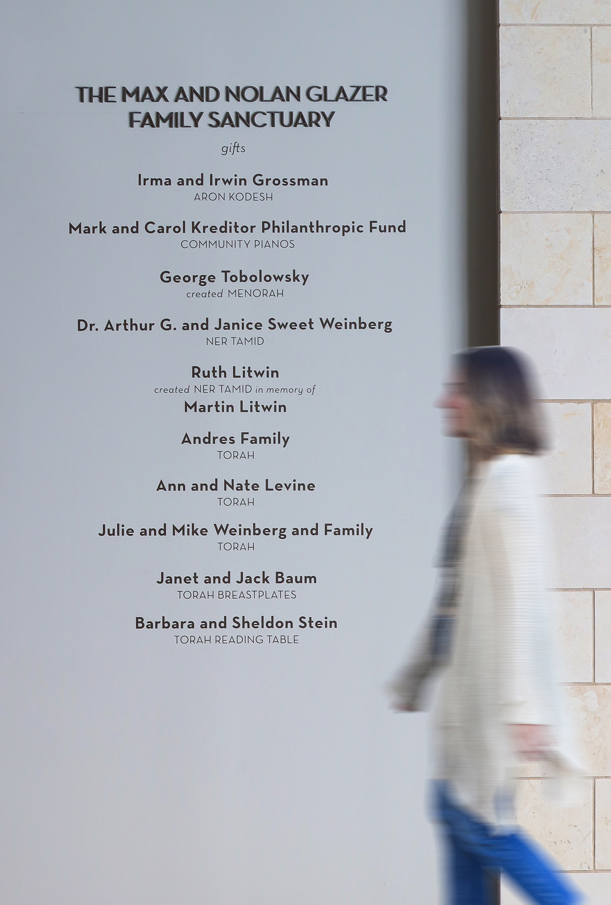 Donor recognition wall reading 'The Max and Nolan Glazer Family Sanctuary' with a list of gift names and dedications including Irma and Irwin Grossman, Ruth Litwin, and Barbara and Sheldon Stein, as a blurred woman walks past a stone-tiled column.