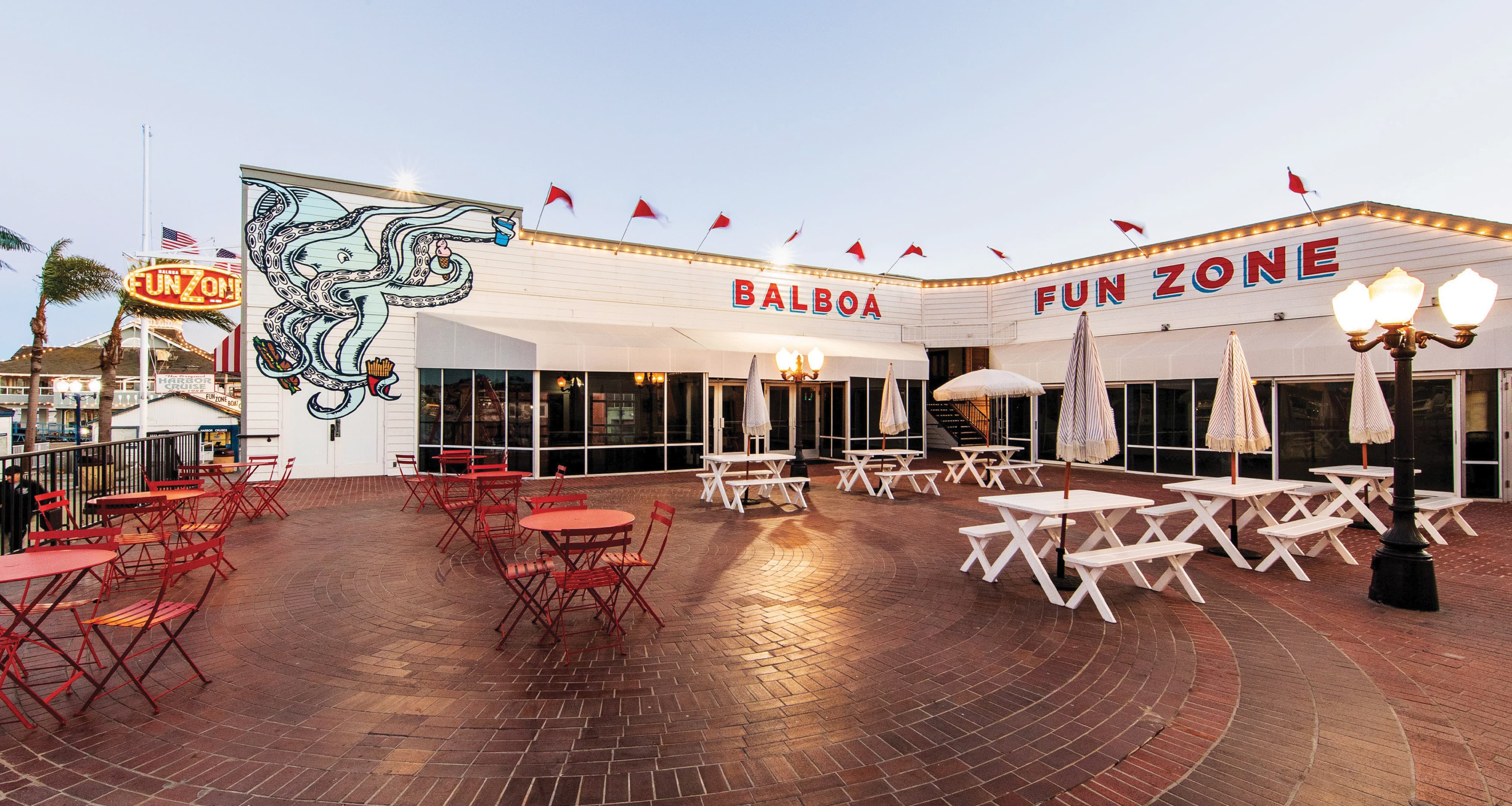 A wide dusk view of the Balboa Fun Zone central courtyard plaza featuring the large-scale "Balboa Fun Zone" roofline identity lettering in red and blue with carnival bulb lighting and red pennant flags, a whimsical illustrated octopus mural on the building corner, red bistro tables and white picnic benches arranged across a circular brick paver plaza, and a classic gas lamp post — demonstrating RSM Design's art programs and public art and murals, identity signage, experiential graphics and branded environments, and placemaking services within the waterfront and mixed-use markets