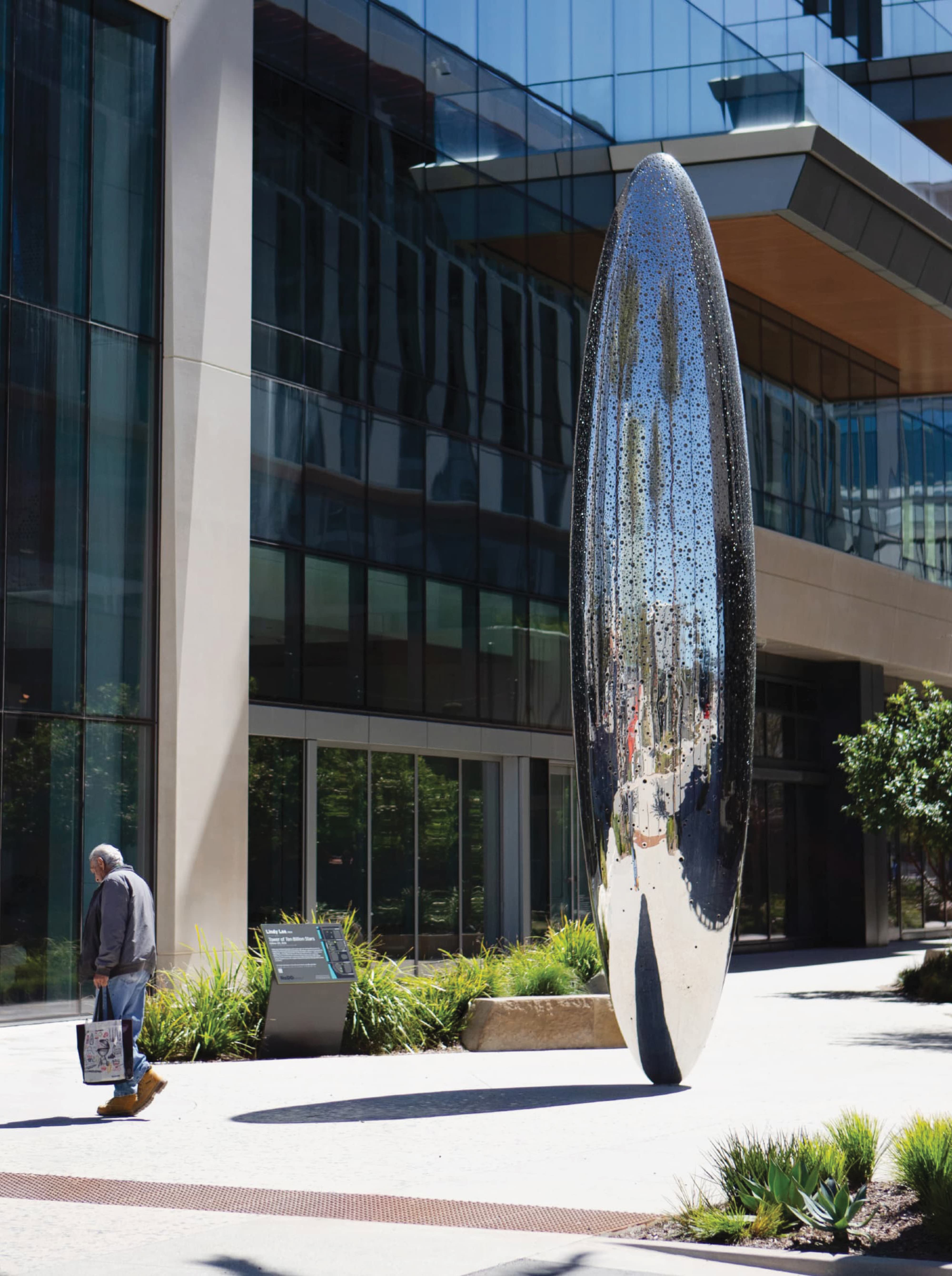 Tall reflective, perforated stainless steel sculpture titled 'Tower of Ten Billion Stars' standing upright on a plaza outside a glass office building, with an interpretive sign beside it and a man walking past carrying tote bags.