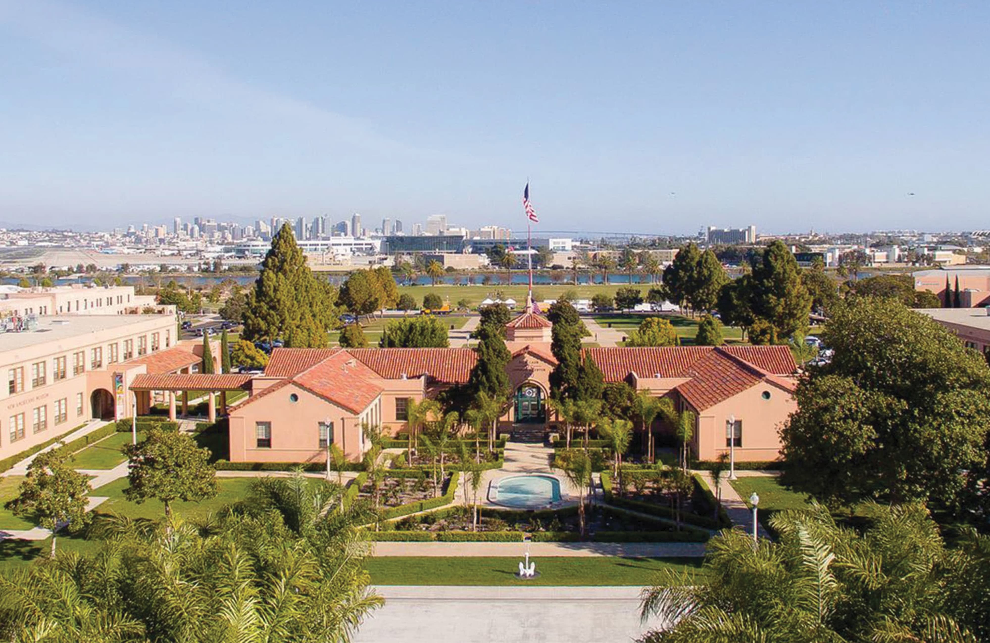 Aerial view of Liberty Station's Spanish-style historic buildings with red tile roofs, landscaped courtyards, a central fountain, and a flagpole, framed by palm trees, with San Diego's skyline and bay visible in the distance.