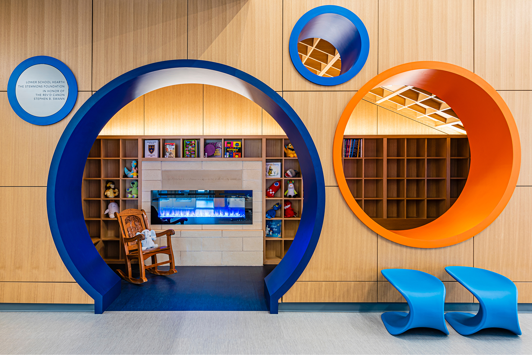 Playful reading nook framed by a large circular blue archway with a fireplace, bookshelves, stuffed animals, and a wooden rocking chair inside, flanked by a smaller blue and a larger orange circular alcove with book storage, blue S-shaped chairs, and a plaque reading 'Lower School Hearth, The Stemmons Foundation, in honor of the Rev'd Canon Stephen B. Swann.