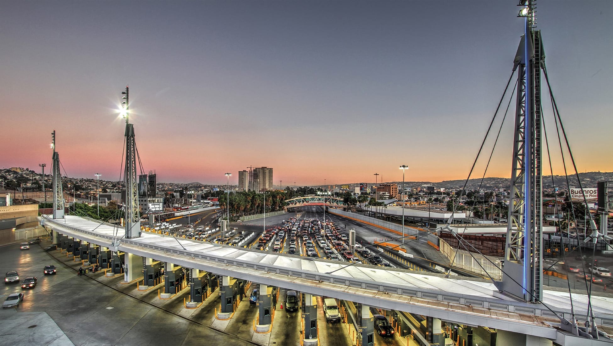 Aerial twilight view of the San Ysidro Port of Entry, showing rows of illuminated vehicle inspection booths beneath a curved canopy with tall cable-supported light towers, long lines of cars waiting to cross, and the Tijuana skyline and hillside city lights in the background at sunset.