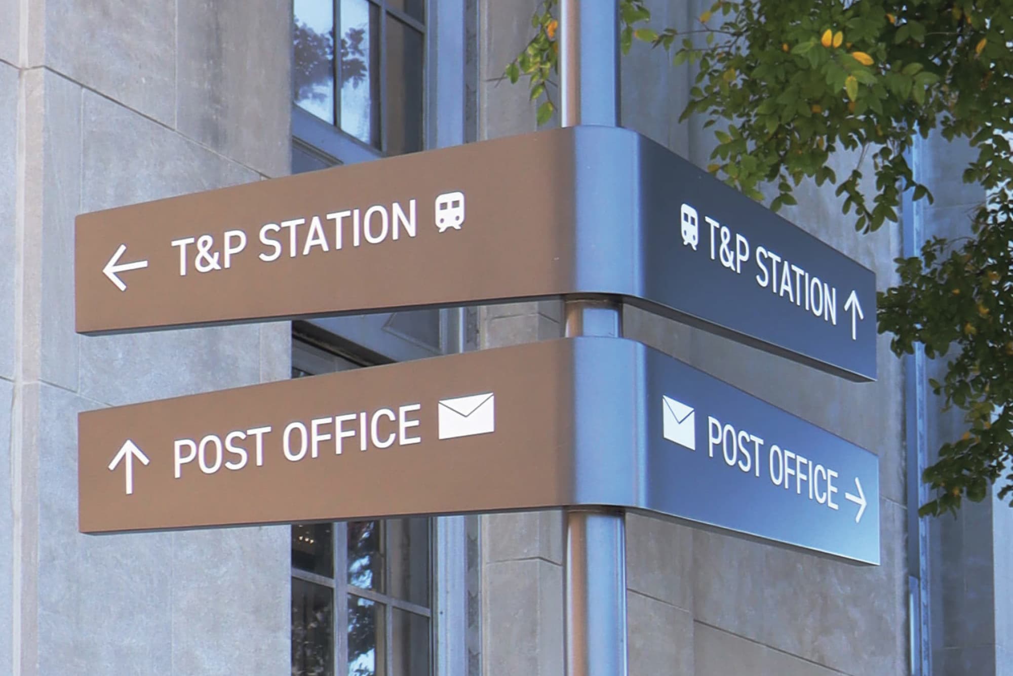 Dual-sided multi-blade directional wayfinding sign in brown and navy directing pedestrians to T&P Station and Post Office with transit and mail icons on a downtown streetscape, showcasing environmental graphic design and signage for a mixed-use transit development by RSM Design.