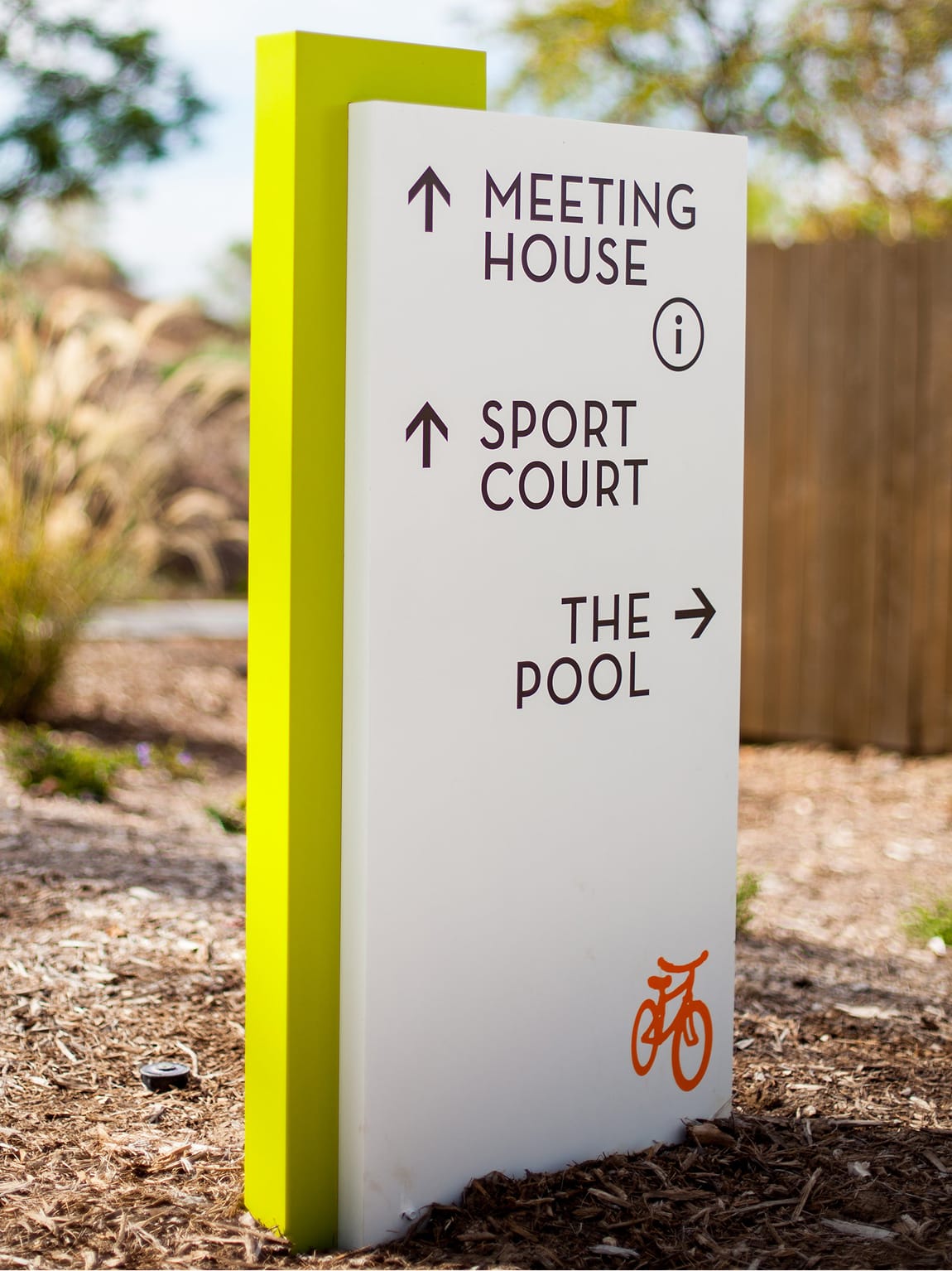 Outdoor wayfinding pylon sign with yellow-green accent post directing visitors to Meeting House, Sport Court, and The Pool with directional arrows and an orange bicycle icon, installed at Great Park Neighborhoods in Irvine, California, designed by RSM Design