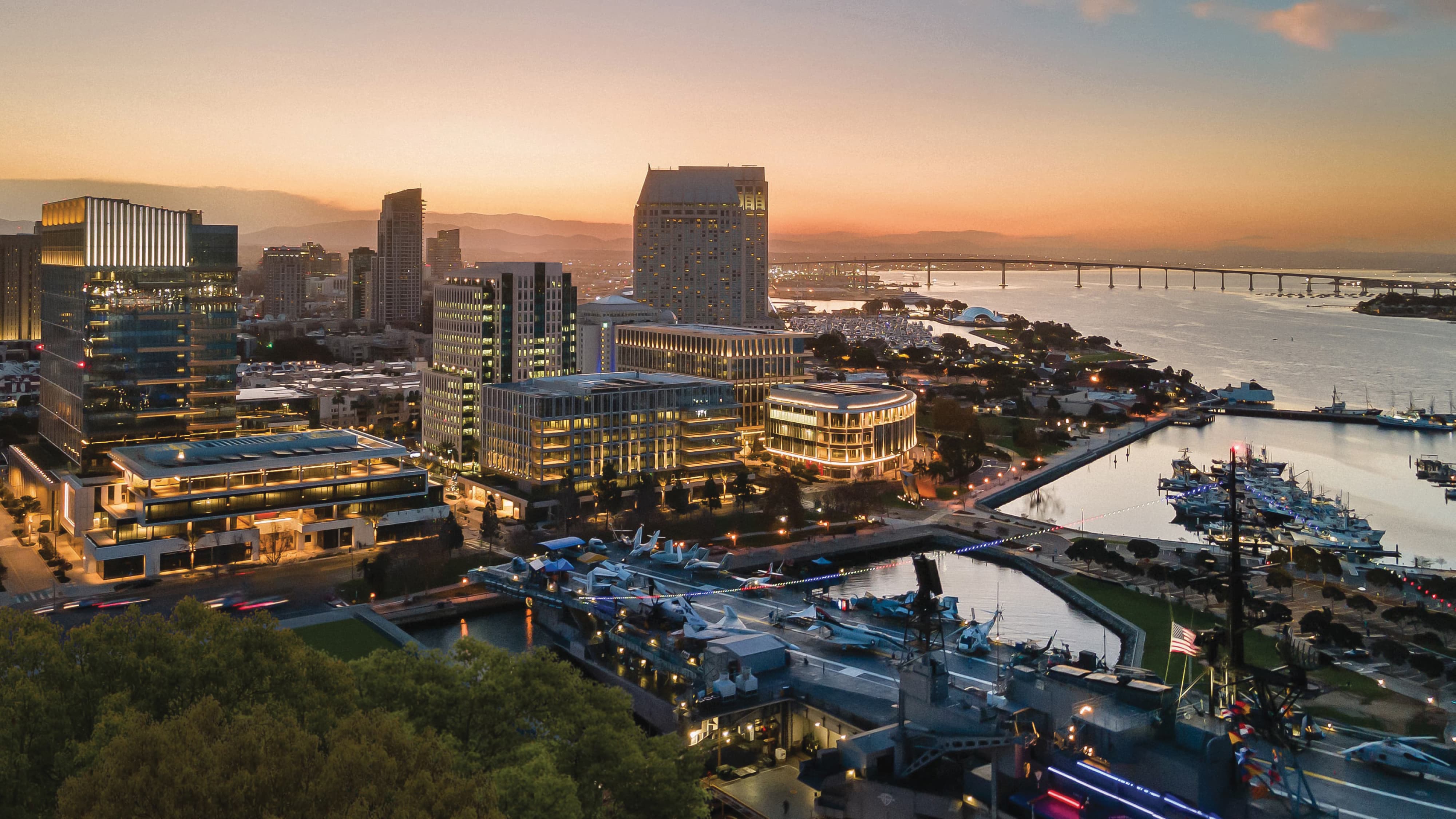 Aerial twilight view of downtown San Diego skyline and waterfront, with illuminated high-rise buildings, the USS Midway Museum aircraft carrier docked in the foreground, a marina, and the Coronado Bridge stretching across the bay under a pink and orange sunset sky.