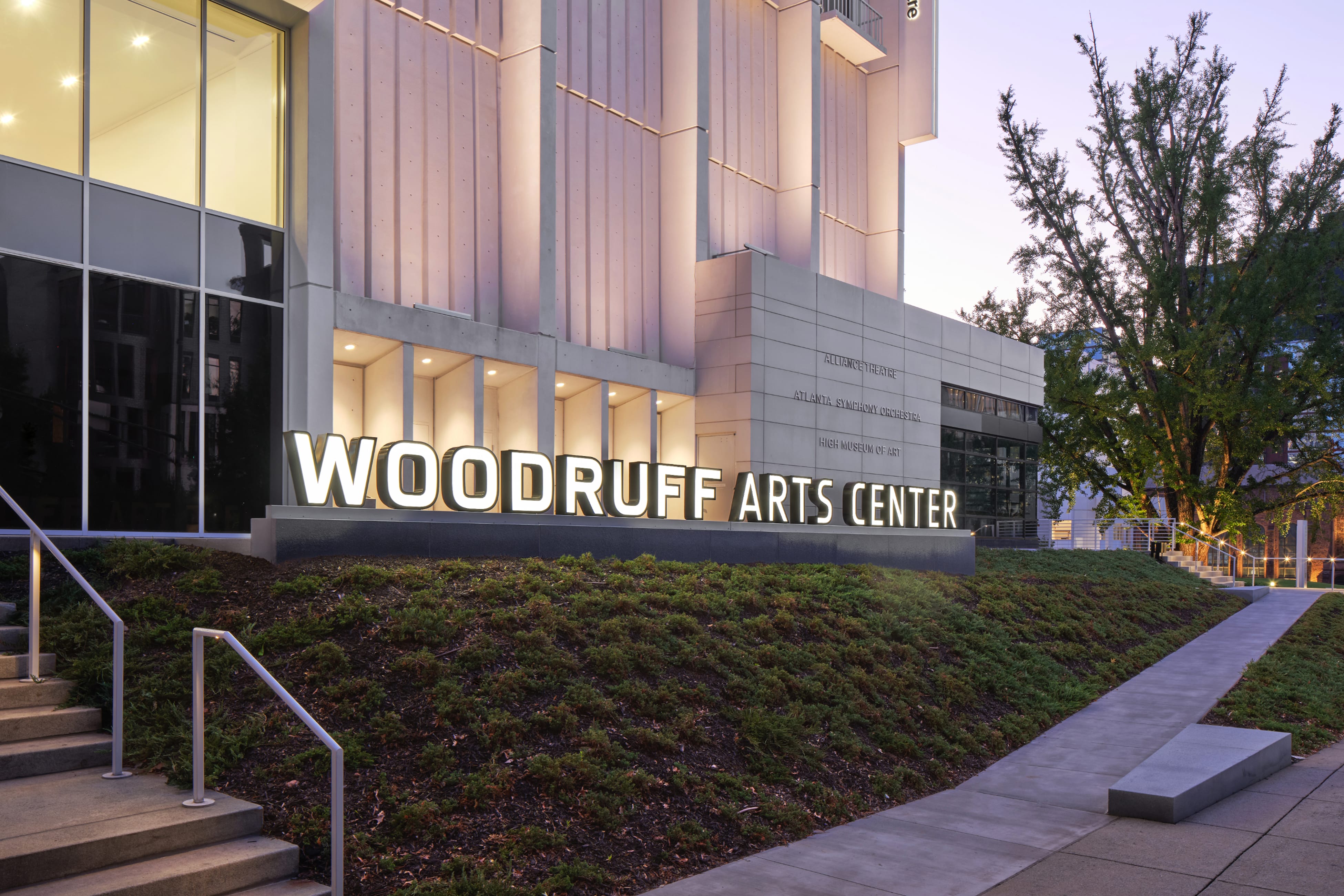 Illuminated "WOODRUFF ARTS CENTER" letters on a landscaped hillside at dusk, with wall-mounted directory listing the Alliance Theatre, Atlanta Symphony Orchestra, and High Museum of Art in the background – campus wayfinding and environmental graphic design by RSM Design.
