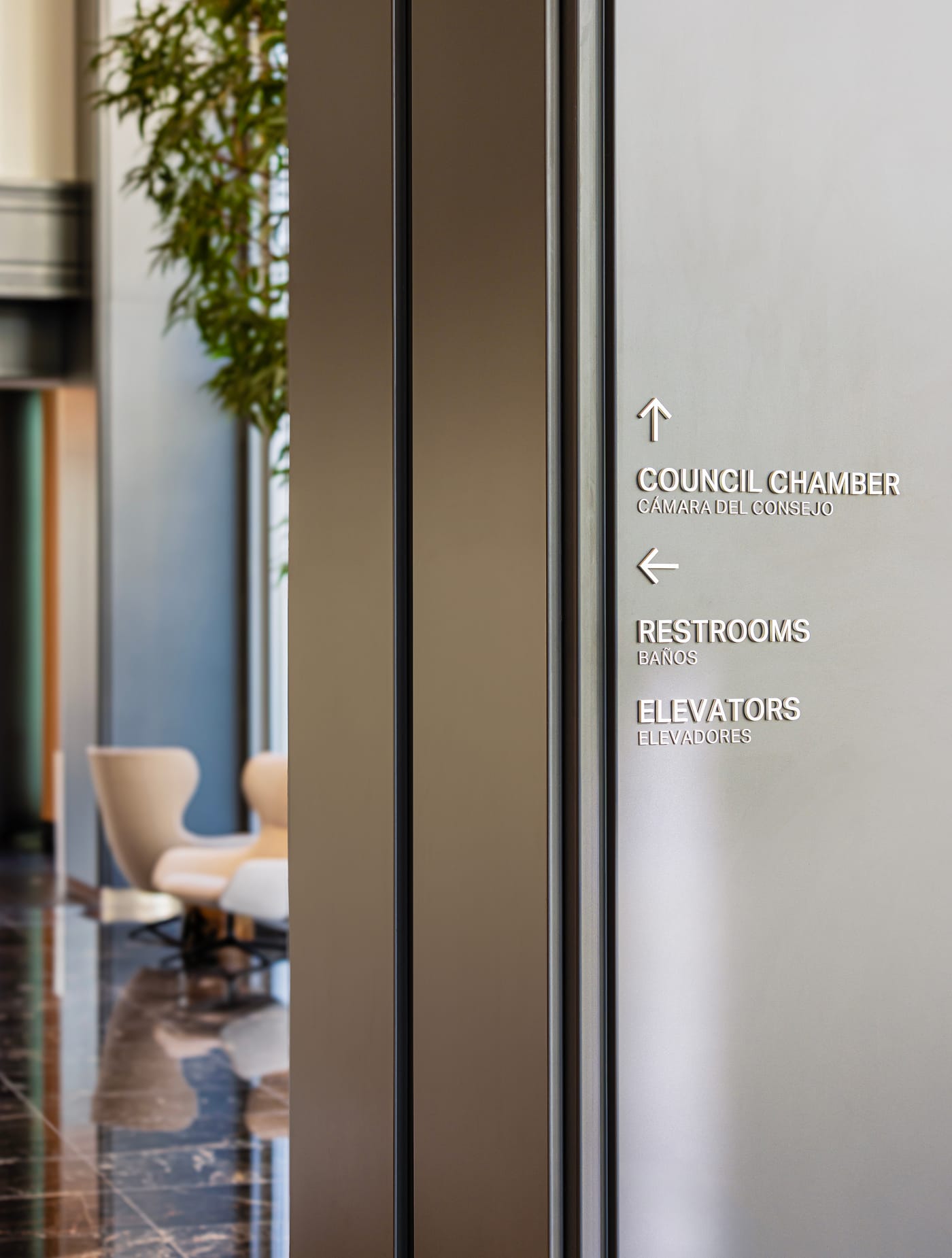 Bilingual interior wayfinding sign in English and Spanish directing visitors to Council Chamber, Restrooms, and Elevators with dimensional white letters on light wall panel at Fort Worth City Hall