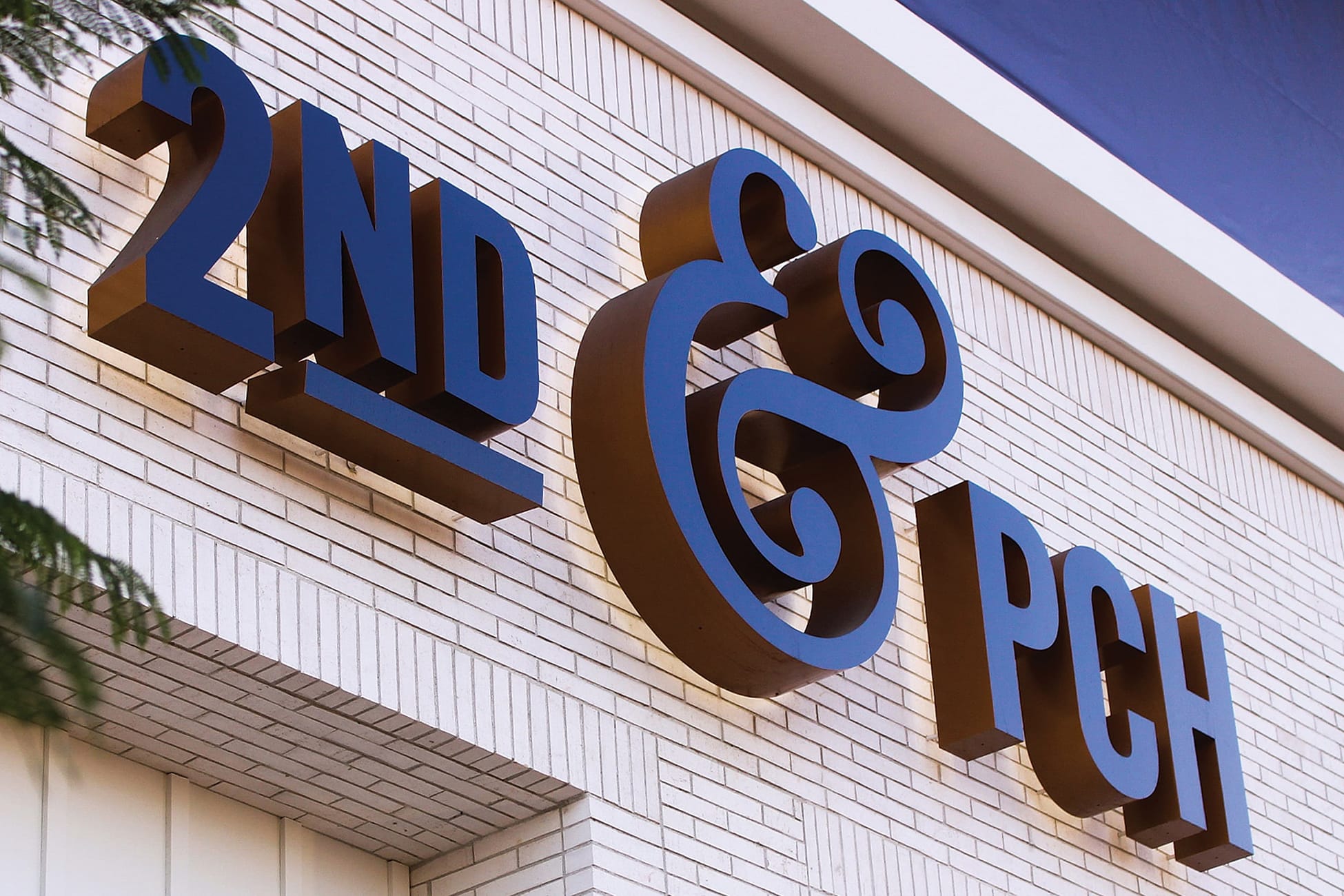 Large-scale dimensional wall-mounted channel letter signage reading '2nd & PCH' in navy and bronze on a white brick building facade in Long Beach, California, featuring an oversized decorative ampersand as an architectural landmark element — showcasing RSM Design's architectural graphics, environmental graphic design, and branded environments for a coastal waterfront retail and mixed-use destination.