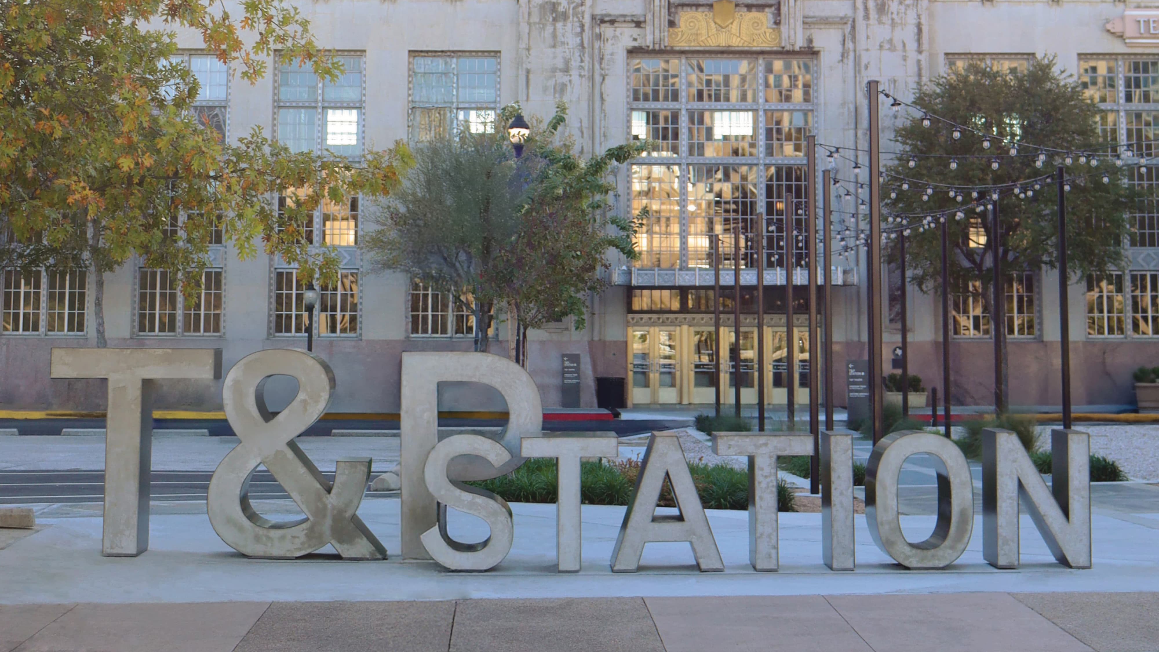 Large-scale concrete dimensional "T&P Station" ground-level letters in an outdoor plaza in front of a historic Art Deco transit building with string lights, showcasing placemaking, experiential graphics, and branded environment design for a mixed-use transit and streetscape development by RSM Design.
